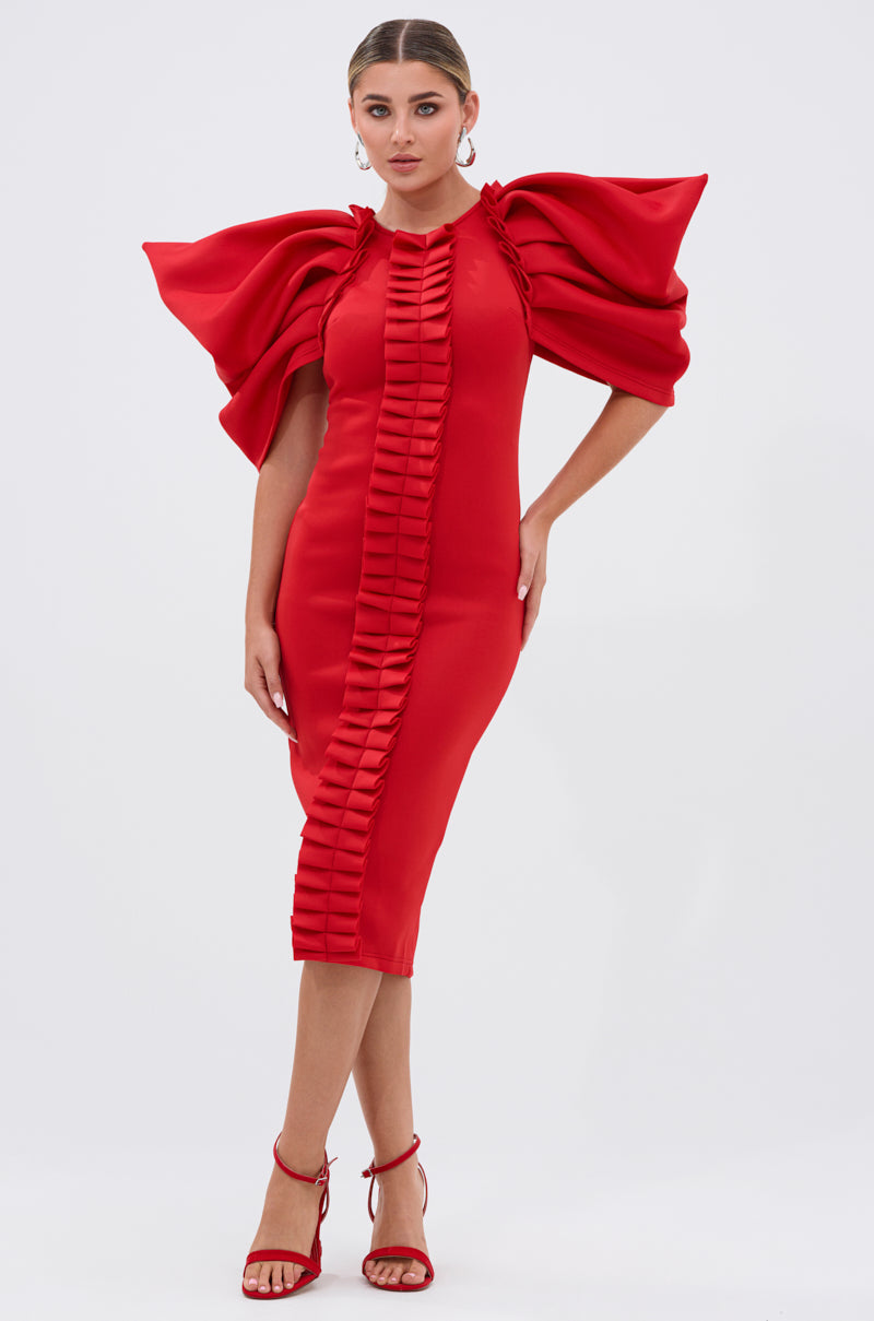 A woman models the PURE JOY MIDI DRESS WITH DRAMATIC SLEEVE IN RED—featuring a fitted silhouette, center pleated ruffle details, and bold sleeves—styled with red heels against a simple light background.