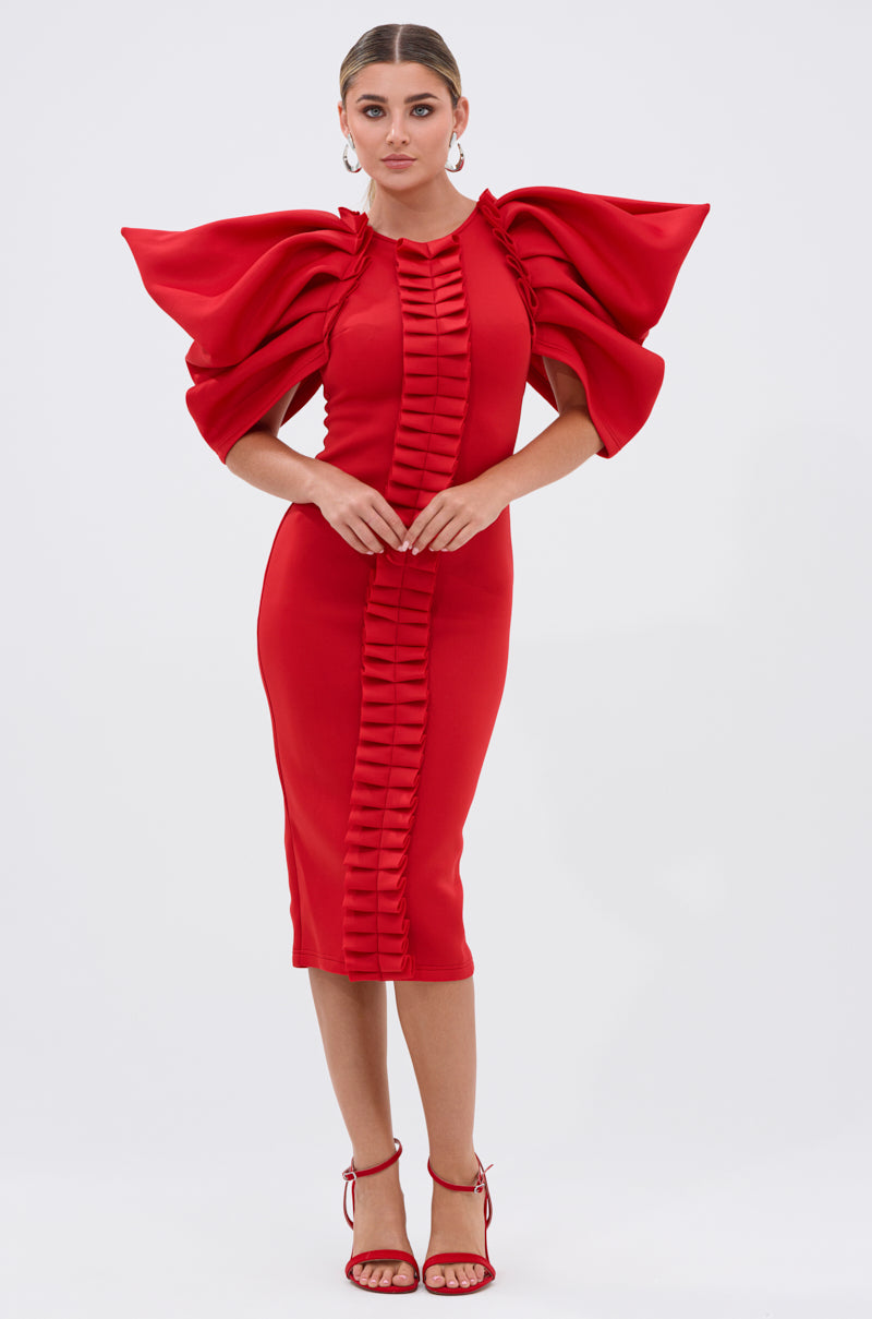 A woman faces forward against a white background in the PURE JOY MIDI DRESS WITH DRAMATIC SLEEVE IN RED, featuring a bodycon fit, bold structured ruffled sleeves, and vertical center ruffle. She pairs it with hoop earrings and red strappy heels; her hair is pulled back.
