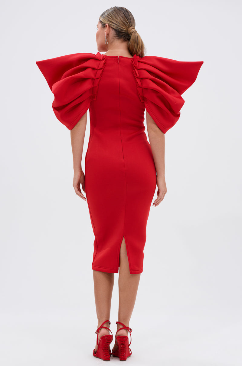 A woman stands facing away in the PURE JOY MIDI DRESS WITH DRAMATIC SLEEVE IN RED, featuring a bodycon fit and back slit. She wears her hair tied back and pairs the dress with matching red heeled sandals.