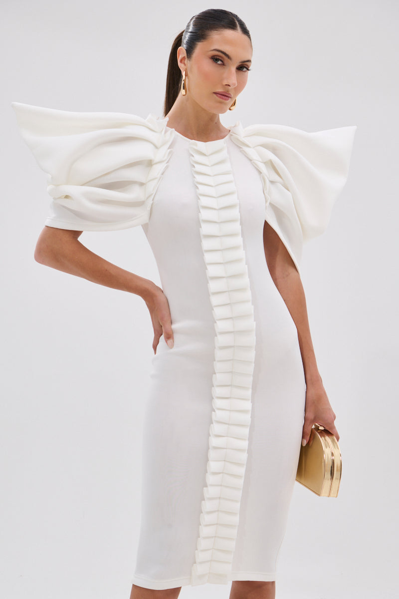 A woman wears the PURE JOY MIDI DRESS WITH DRAMATIC SLEEVE IN WHITE, featuring sculptural ruffled sleeves and vertical ruffles in front. She holds a small gold clutch and poses against a plain light background.