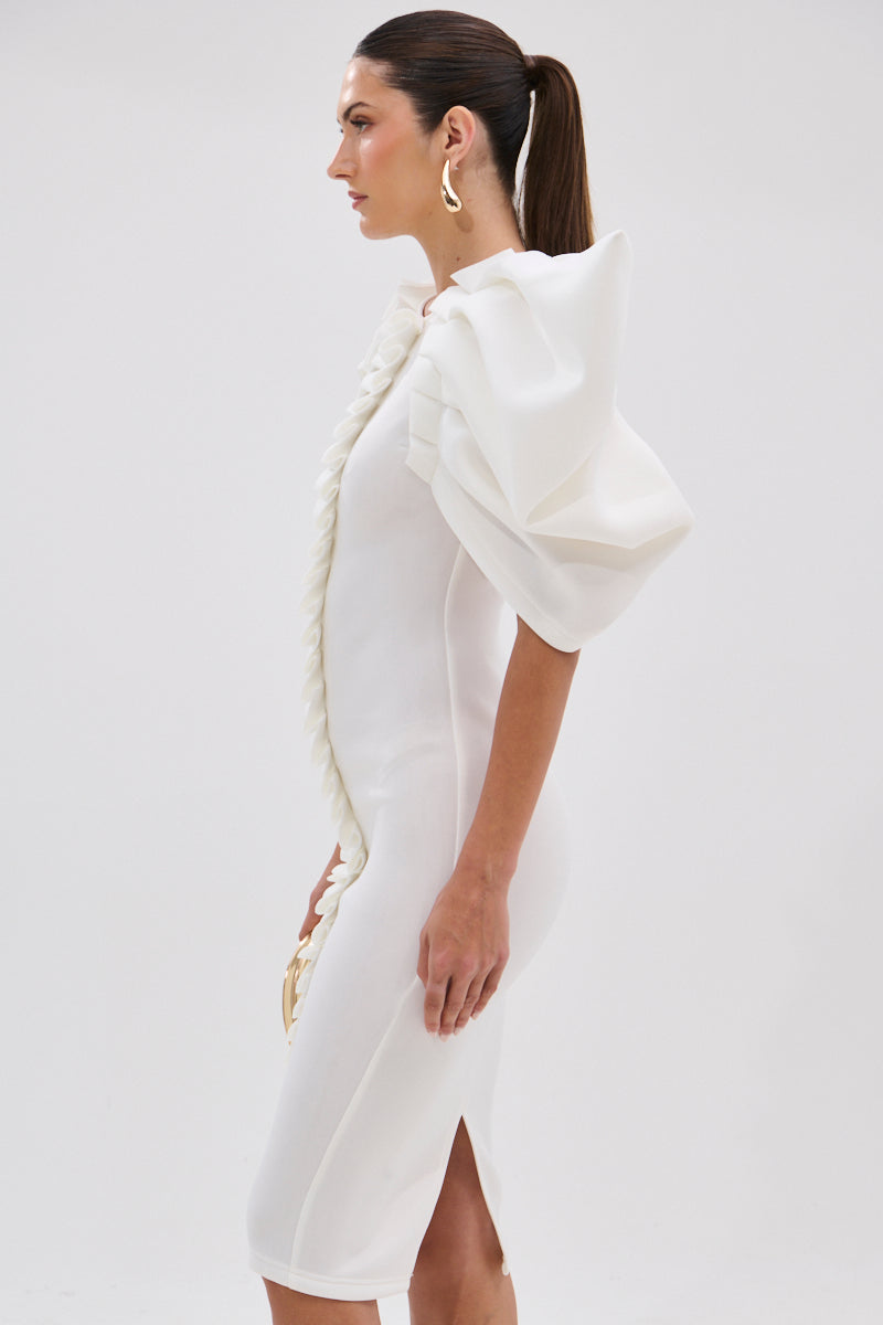 A woman with long hair tied back and large gold earrings poses in profile, wearing the PURE JOY MIDI DRESS WITH DRAMATIC SLEEVE IN WHITE. The fitted dress features puffed sleeves, a side slit, and a decorative front ruffle.