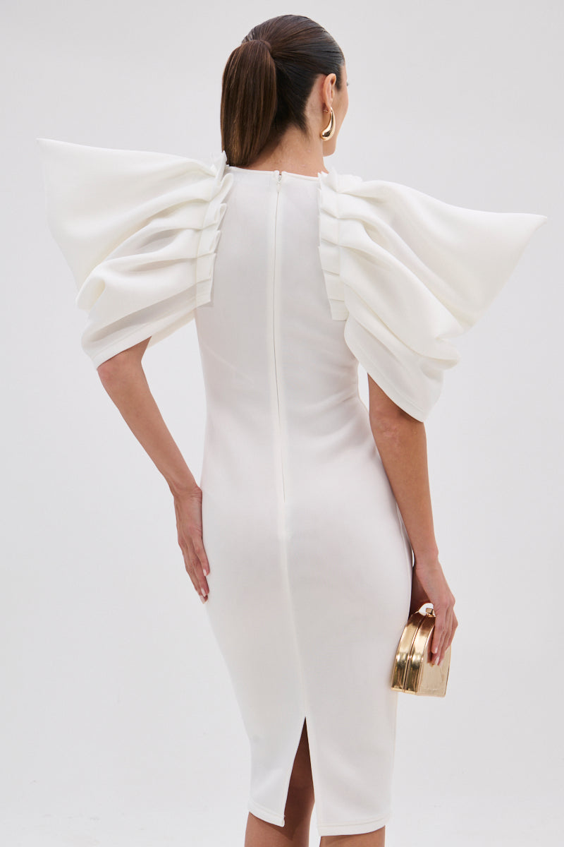 A woman in the PURE JOY MIDI DRESS WITH DRAMATIC SLEEVE IN WHITE stands facing away, holding a gold clutch. Her hair is styled in a low ponytail and she accessorizes with large gold hoop earrings.