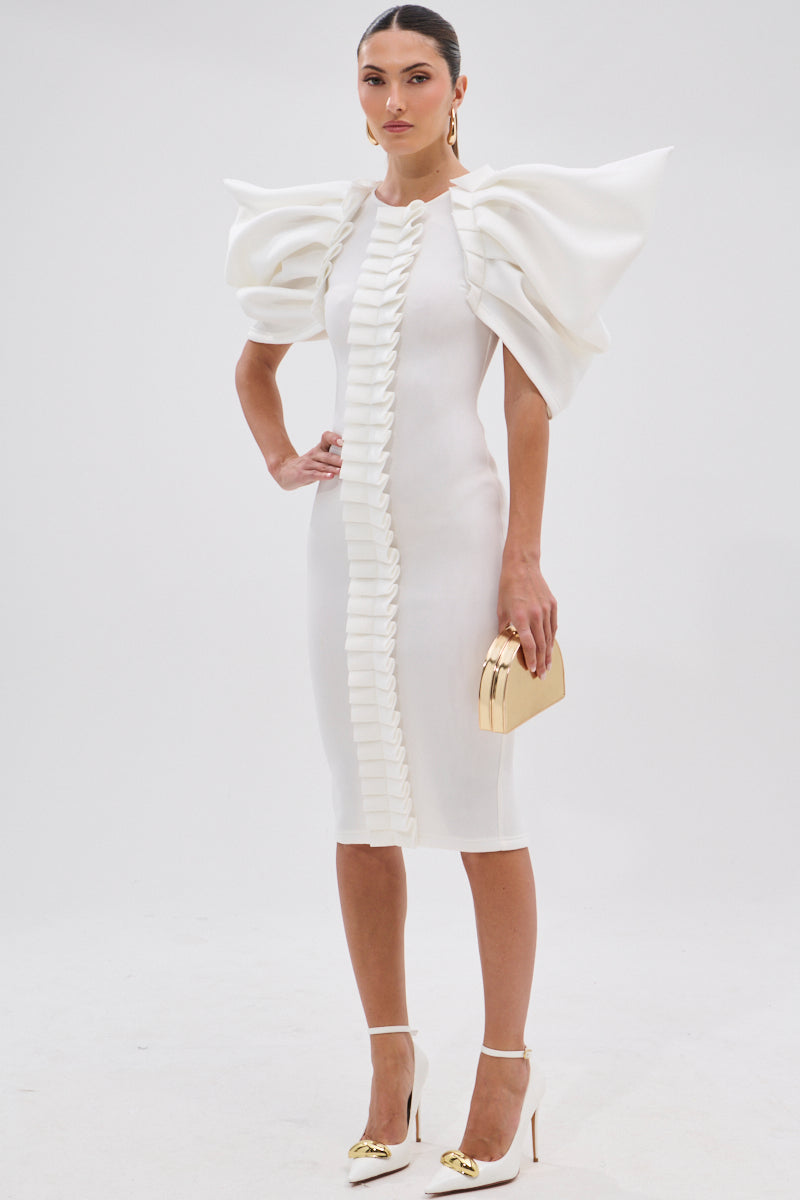 A woman wears the PURE JOY MIDI DRESS WITH DRAMATIC SLEEVE IN WHITE, featuring oversized ruffled sleeves and vertical ruffle accents. She stands against a plain background, holding a gold clutch and wearing white heels with gold accents.