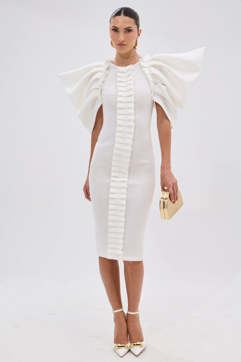 A woman poses in the PURE JOY MIDI DRESS WITH DRAMATIC SLEEVE IN WHITE, featuring sculptural ruffled sleeves and vertical ruffle details. She accessorizes with a gold clutch and gold pointed heels against a plain white background.