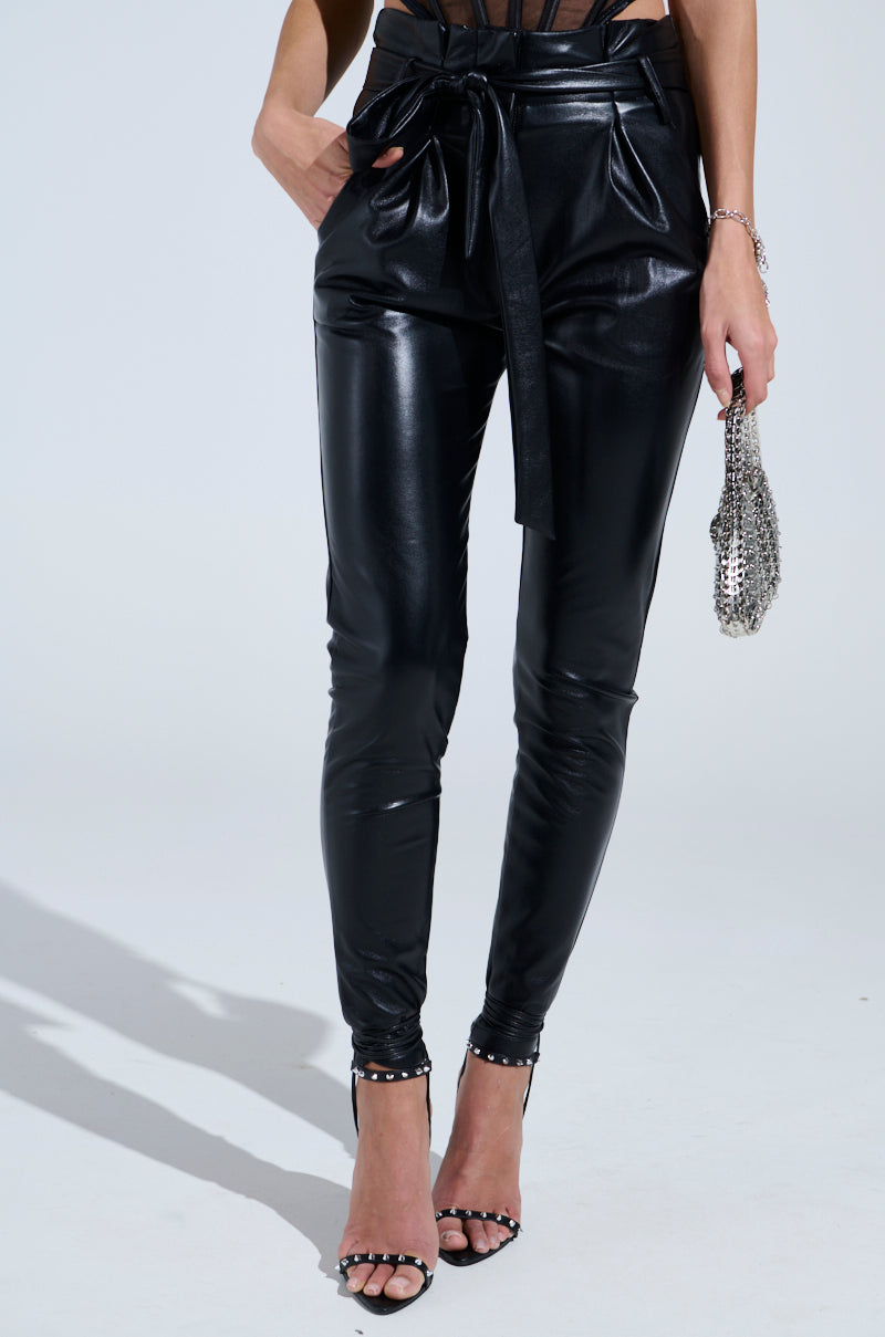A person showcases the LILA PAPERBAG FAUX LEATHER PANTS with a tapered fit and matching tie belt, holding a small chainmail purse and wearing black open-toe heels against a plain white backdrop.