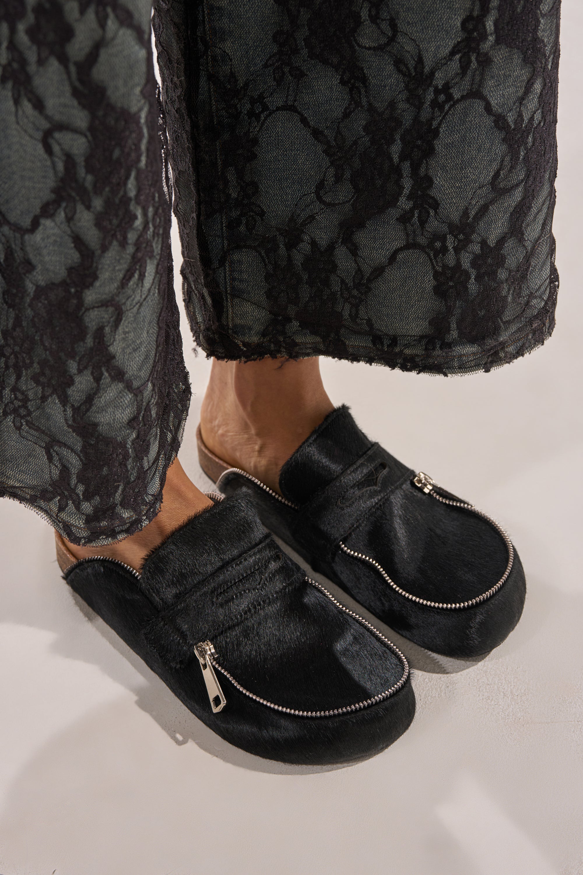 Close-up of a person wearing AZALEA WANG LIMERICK BLACK METAL EMBELLISHMENT FLAT—black slip-on flats with zippers, faux fur texture, metal details, and cushioned footbed—paired with dark lace-trimmed pants. 0
