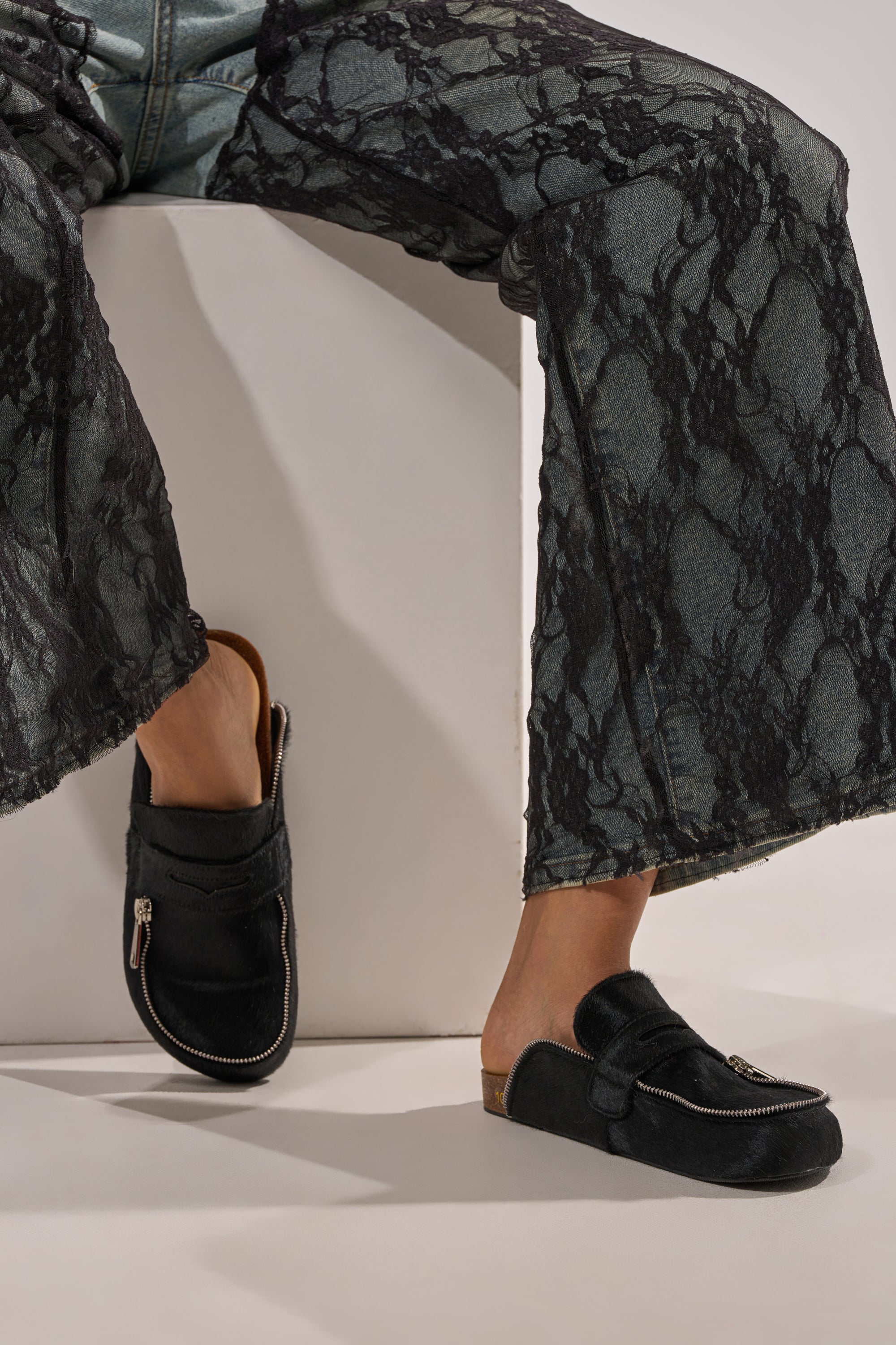 A person wearing black lace overlay pants and AZALEA WANG LIMERICK BLACK METAL EMBELLISHMENT FLAT mules sits on a white block, one foot flat on the ground and the other leg bent. 1