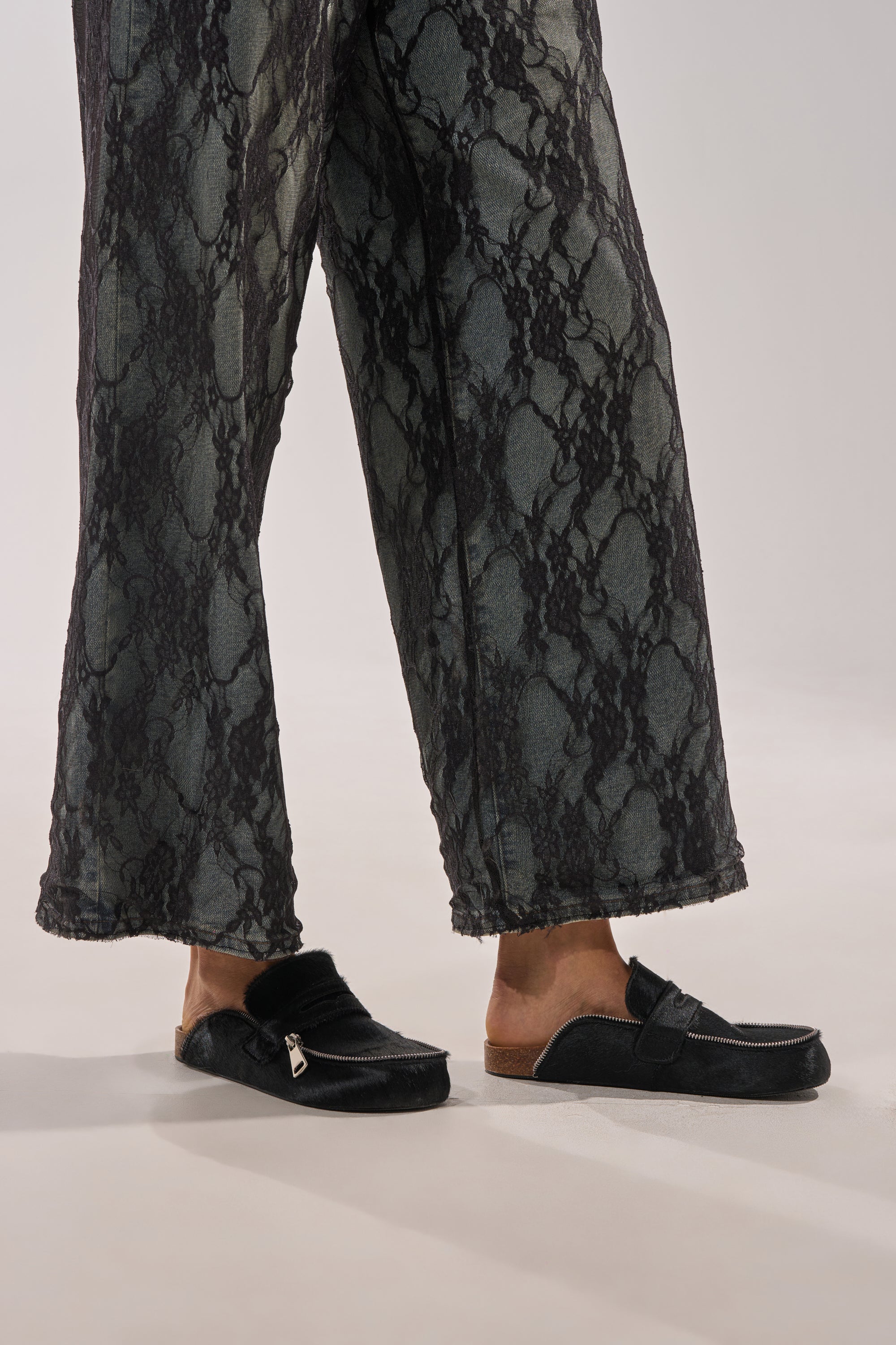 A person wearing snake-print pants and AZALEA WANG LIMERICK BLACK METAL EMBELLISHMENT FLAT mules stands on a light surface with feet slightly apart, facing left. 2