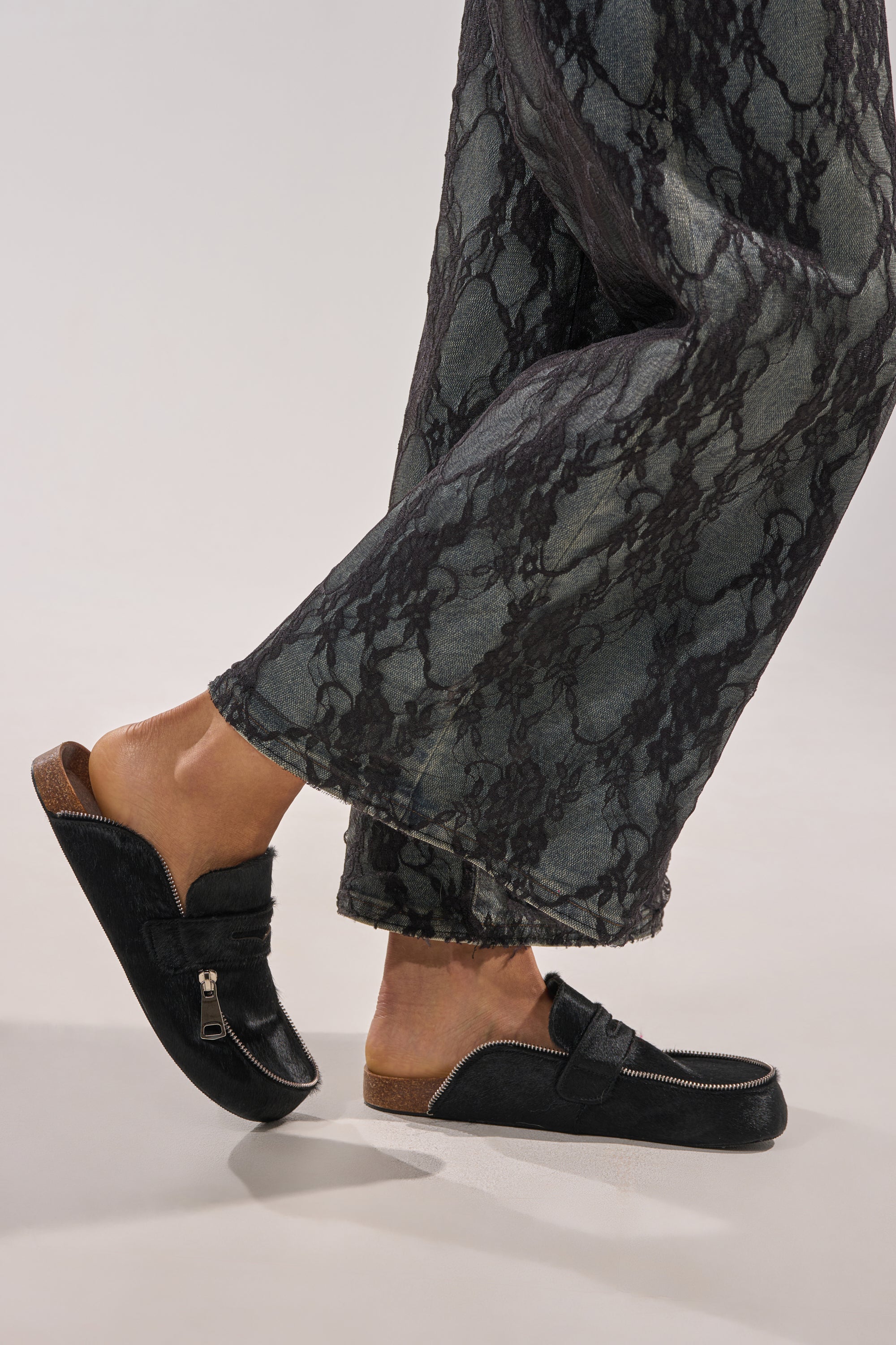 A person wears the AZALEA WANG LIMERICK BLACK METAL EMBELLISHMENT FLAT loafers with zippers and a cushioned footbed, paired with wide-leg lace pants, standing on a light-colored surface. 7