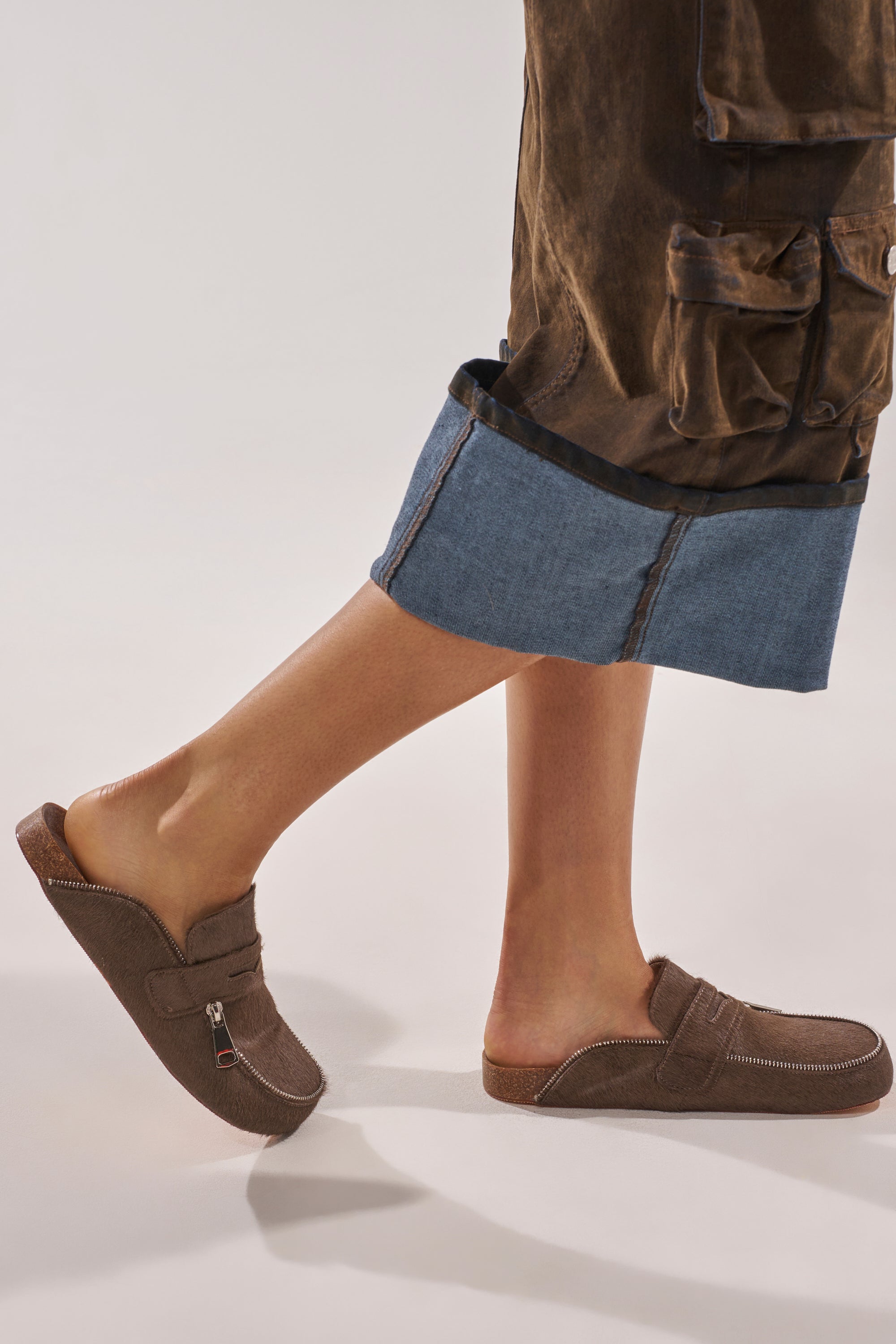 A person standing on tiptoe against a neutral background, wearing cropped denim pants with brown pockets and the AZALEA WANG LIMERICK CHOCOLATE METAL EMBELLISHMENT FLAT loafers. 1