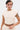 A person with short black and blonde hair wears the SYDNEY TEXTURED BLOUSE—cream-colored, sleeveless, ruched, and cinched—with matching pants. They show tattoos, hoop earrings, and nose and navel piercings against a plain white background.