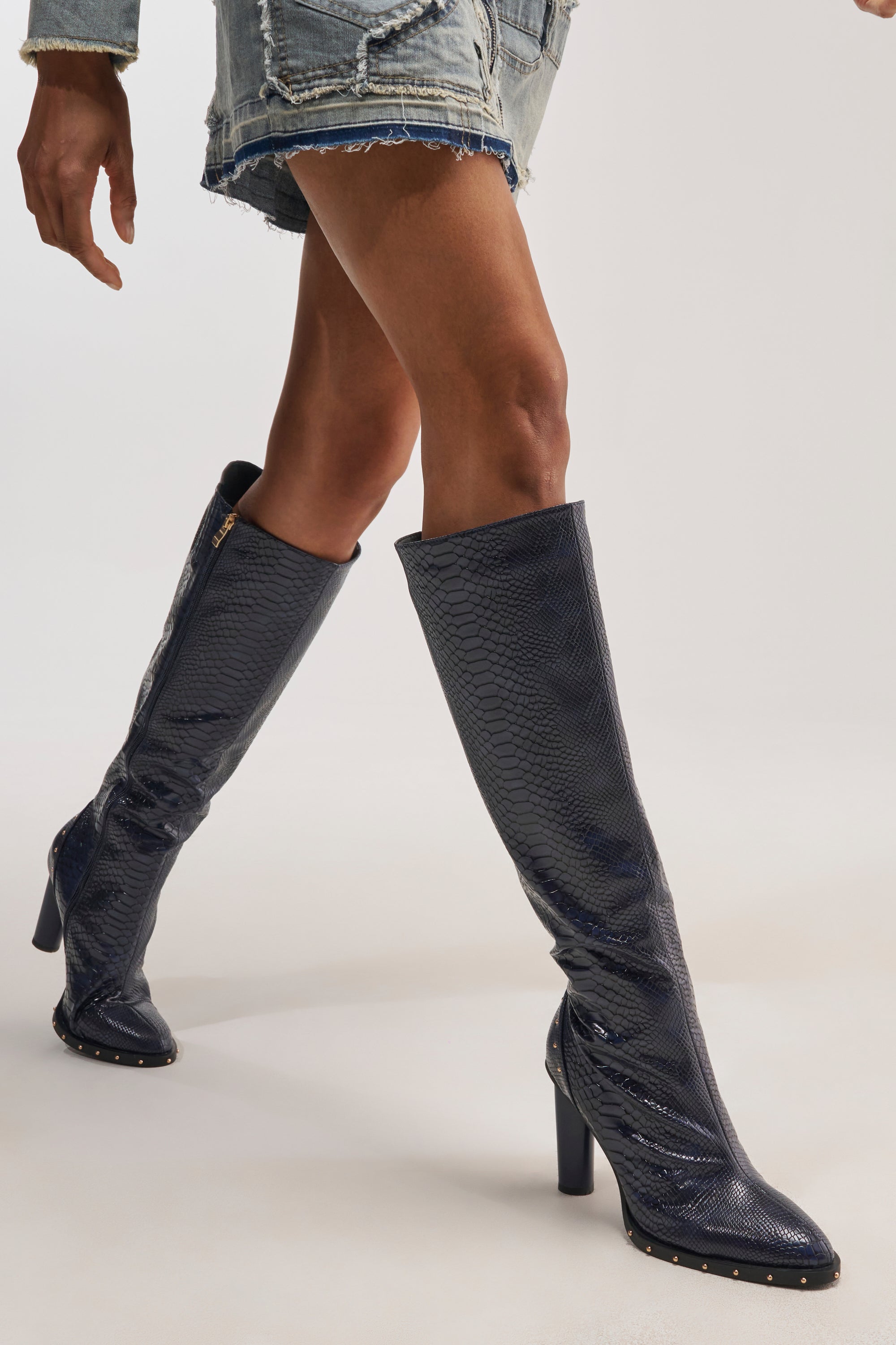 A person wearing the AZALEA WANG LIOREN NAVY SNAKE BLOCK KNEE HIGH BOOT—knee-high boots in a navy snake-print design crafted from snake-embossed faux leather, featuring high heels and studded soles—paired with distressed denim shorts, walking against a plain background.