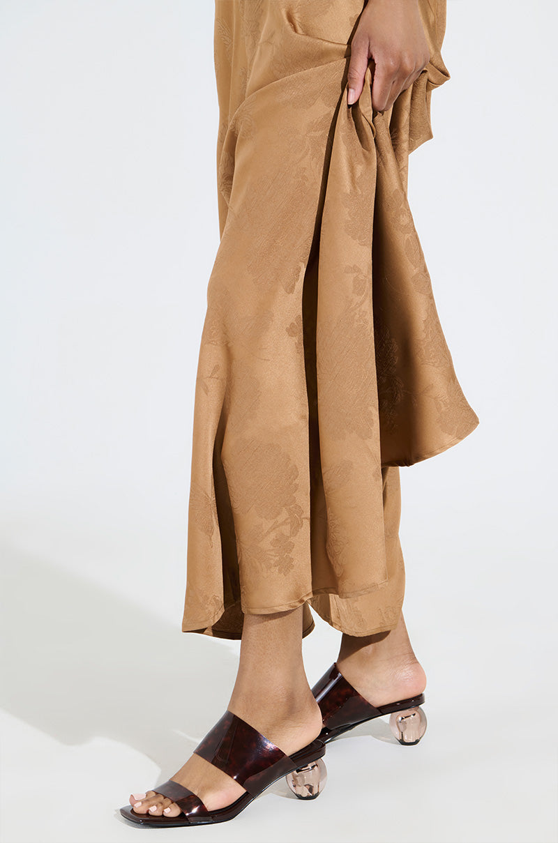 A person models a patterned tan outfit while holding one side, highlighting the AZALEA WANG LIRIO BROWN BALL HEEL SANDAL with an open square toe and transparent novelty heels, against a plain white background.