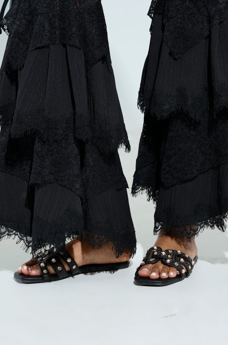 A person stands against a light gray background wearing layered black lace pants and LISA BLACK EMBELLISHED SLIP ON SANDALS with an open square toe and chunky heel.