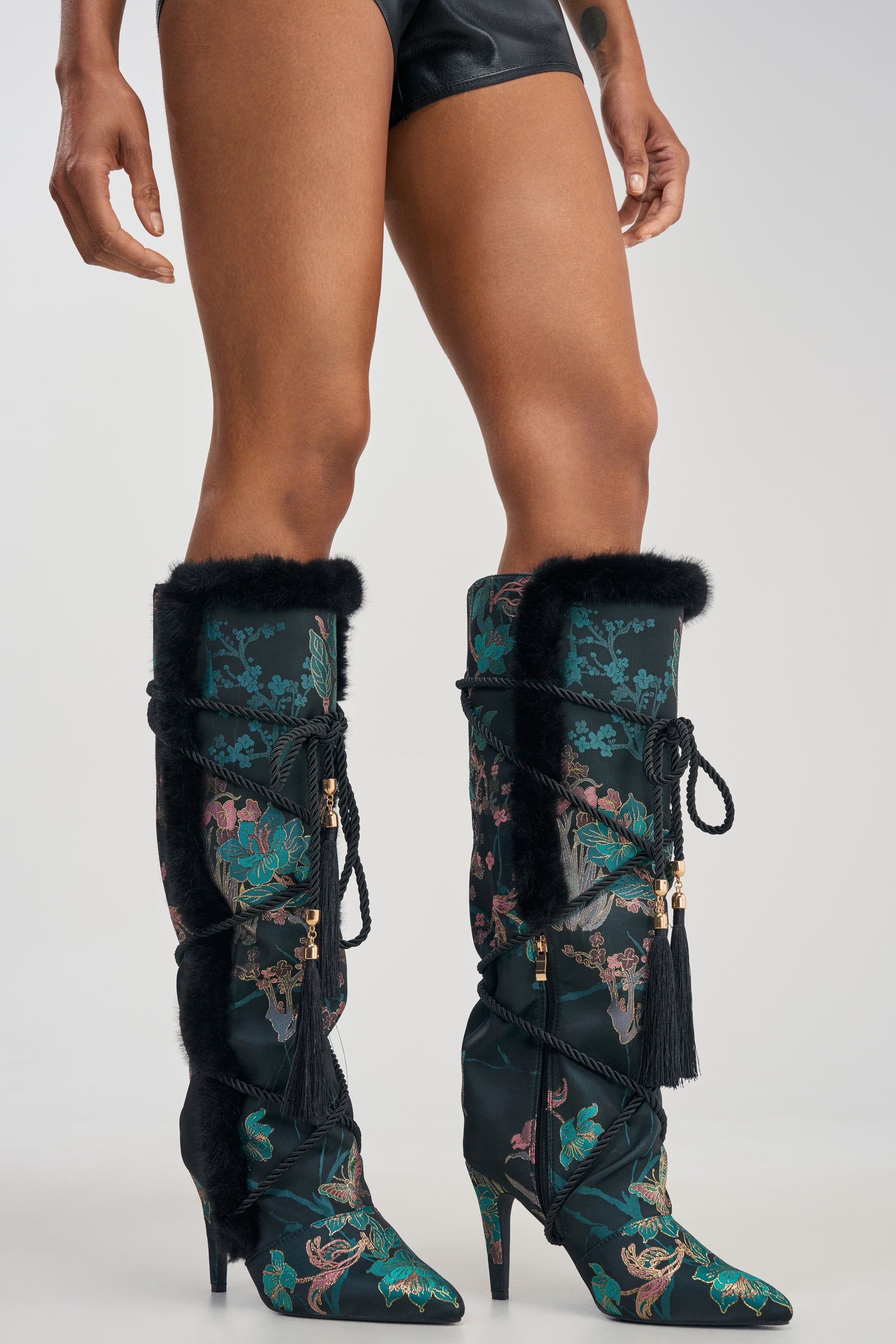 A person wearing the AZALEA WANG LISBETH BLACK MULTI BROCADE AND FUR STILETTO BOOT, styled with black leather shorts.