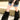Only the lower half of a person is visible as they walk outdoors on a gravel path in a black patterned shirt, denim mini skirt, and AZALEA WANG LISBOA BLACK FUR FOLD OVER BOOTS with chunky heels next to a concrete barrier.