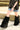Only the lower half of a person is visible as they walk outdoors on a gravel path in a black patterned shirt, denim mini skirt, and AZALEA WANG LISBOA BLACK FUR FOLD OVER BOOTS with chunky heels next to a concrete barrier.