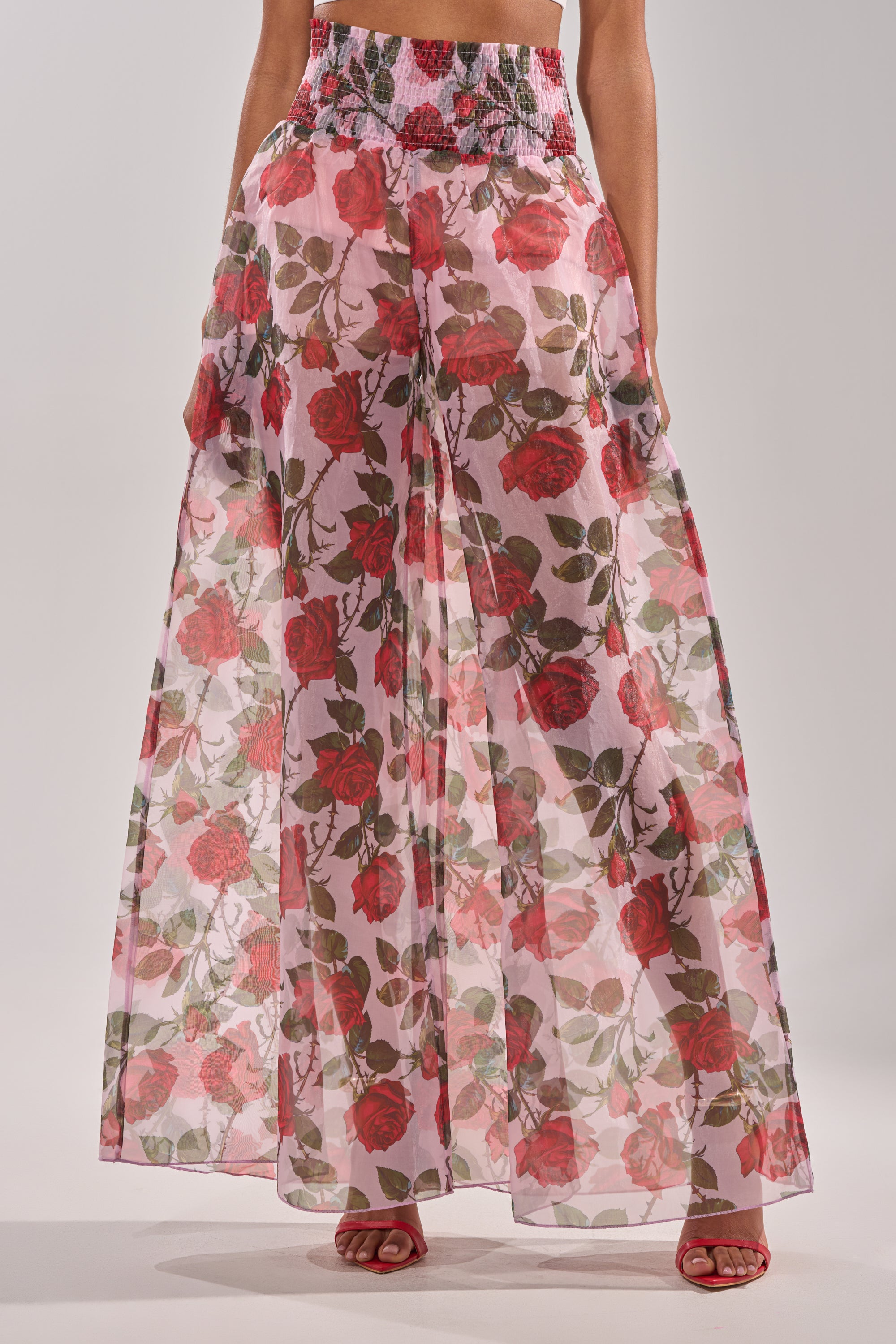 A person wearing the PRETTY GIRL FLORAL TROUSER, featuring a red rose print on a pink base with a high-rise waistband, styled with red heeled sandals. 0