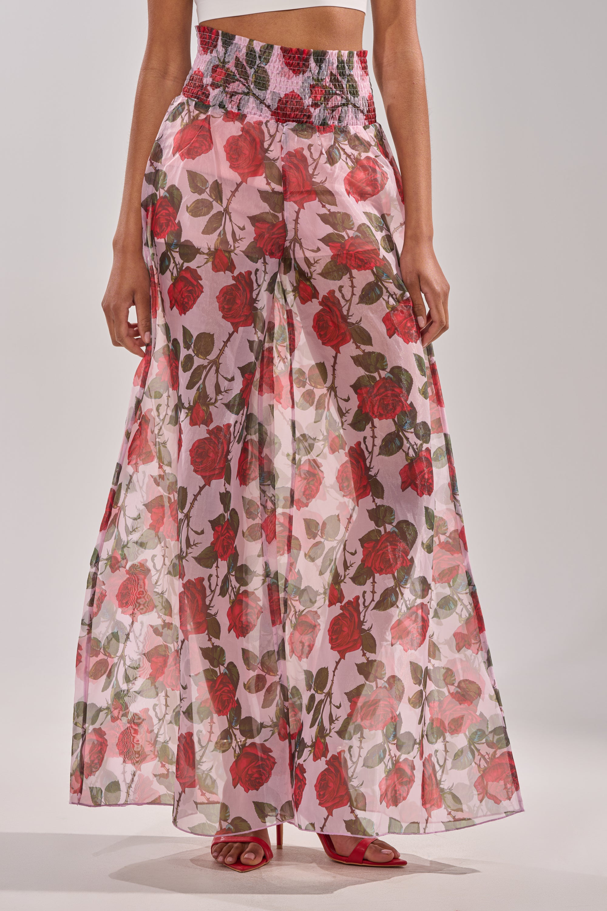 A person models the PRETTY GIRL FLORAL TROUSER—sheer, rose-printed, wide-leg pants with a high-rise smocked waistband—paired with red high-heeled sandals. The image is cropped from midriff to feet. 1