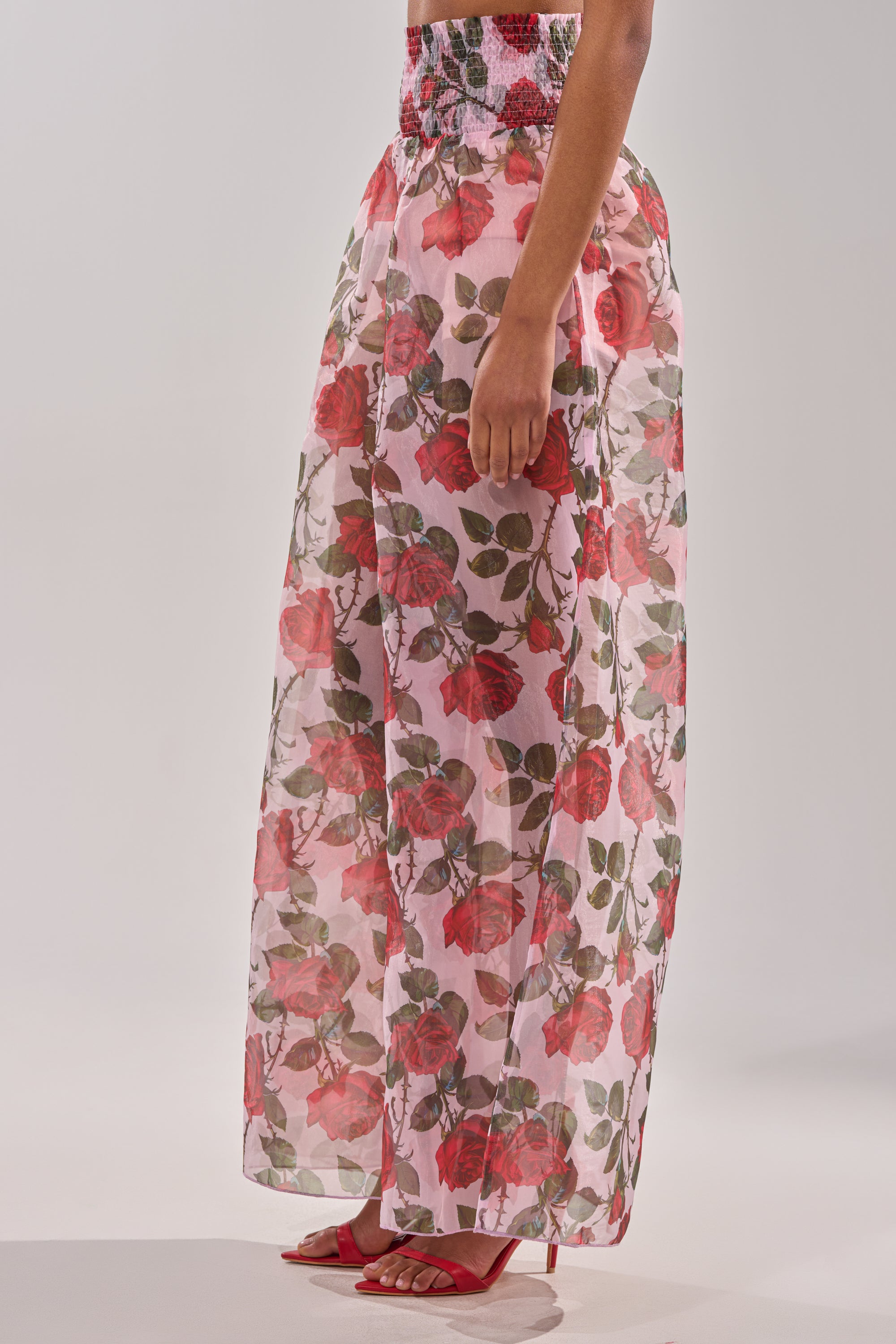A person wears the PRETTY GIRL FLORAL TROUSER with a vibrant red and green floral pattern, reminiscent of a sheer ankle-length skirt, paired with red open-toe high heels against a plain light background. 2