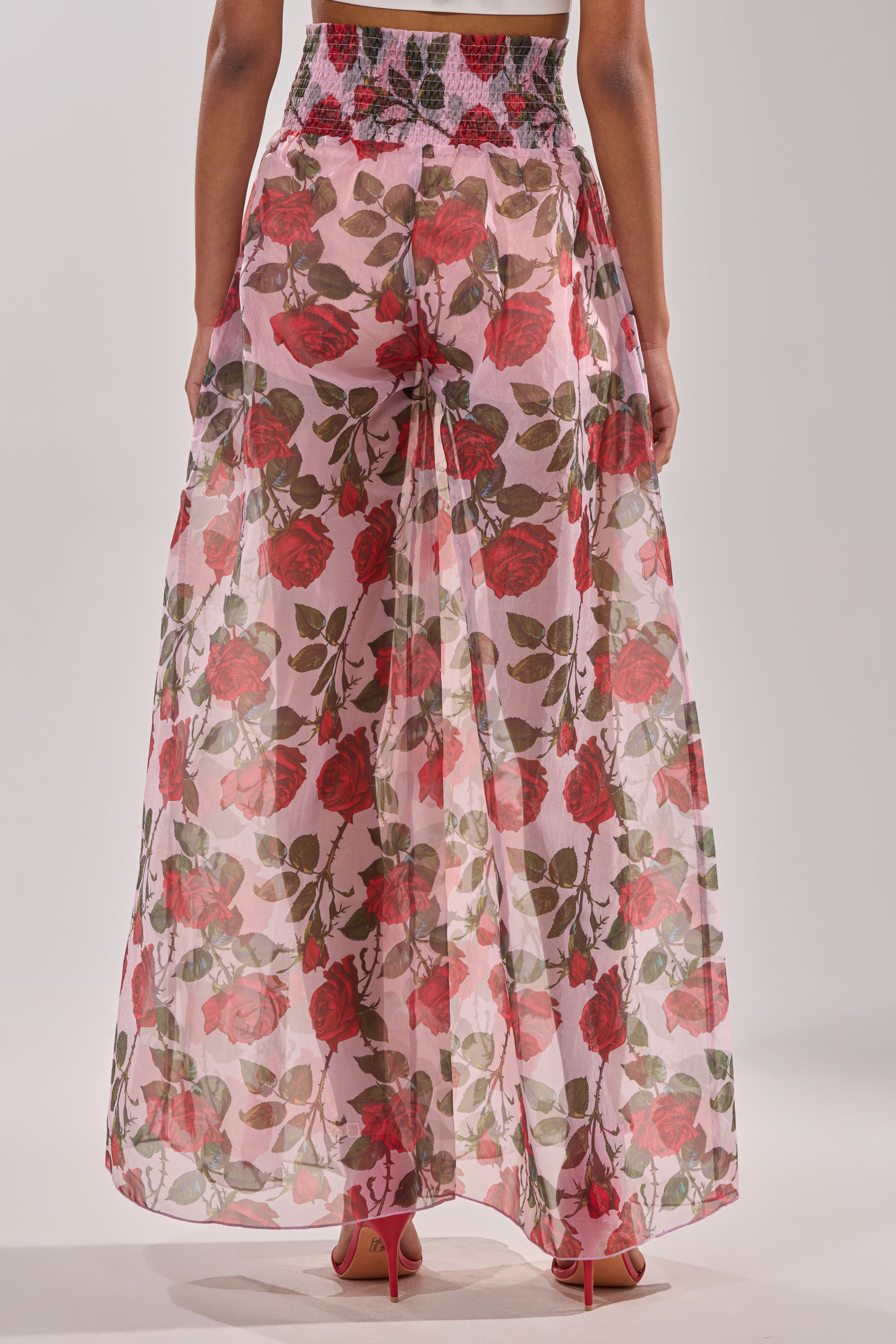 A person faces away, wearing the PRETTY GIRL FLORAL TROUSER—a sheer pink pant with a red rose floral pattern and high-rise waist, partially revealing pink heels underneath. 3