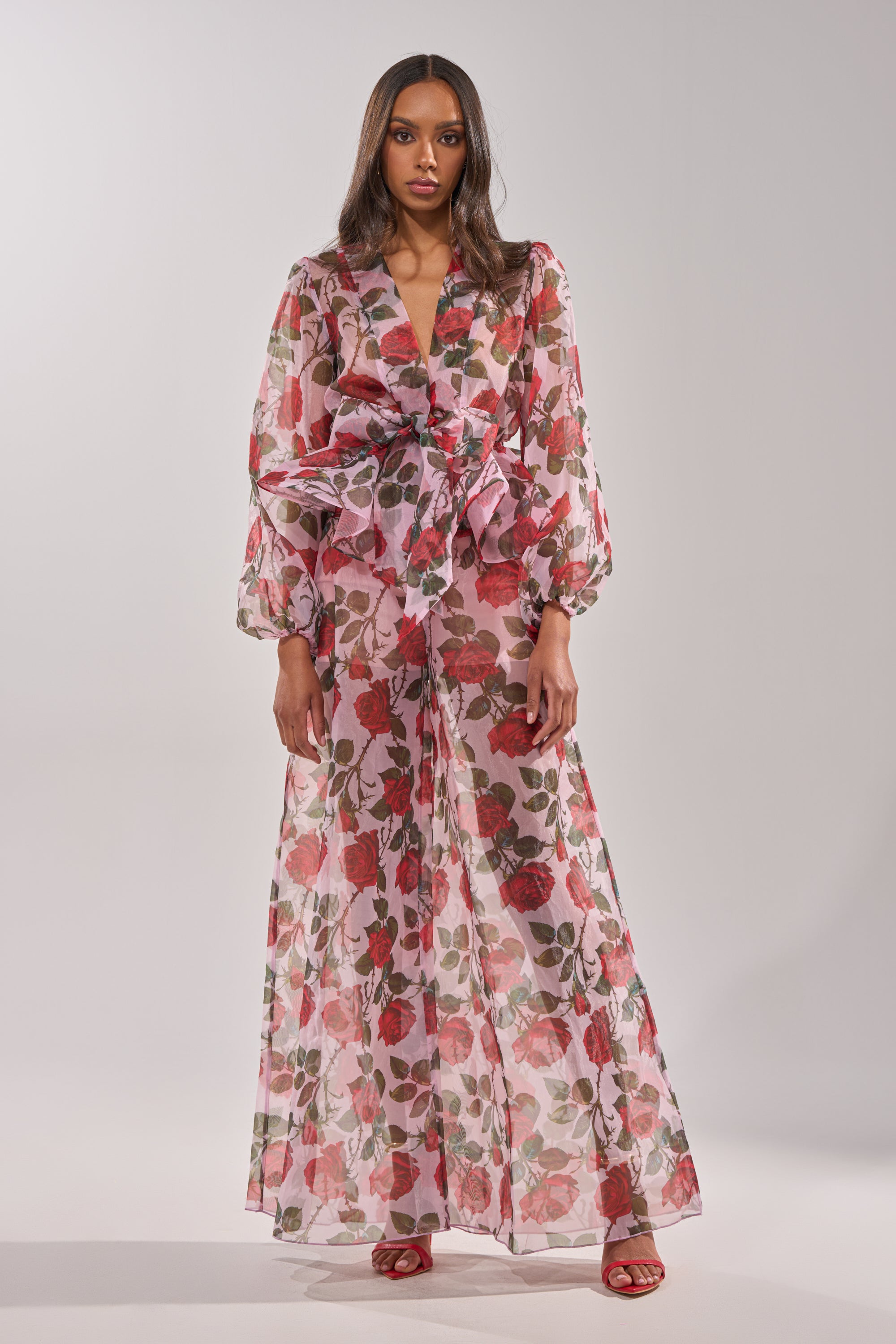 A woman wears PRETTY GIRL FLORAL TROUSER—a sheer, long-sleeved pink, red, and green rose print jumpsuit with high-rise bow waistband and wide legs—paired with red heeled sandals against a plain light gray background. 4