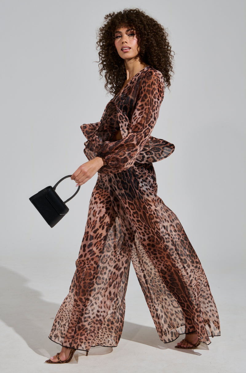 A person with curly hair wears the CHEETAH GIRL PALAZZO PANT, posing confidently in the sheer cheetah-print outfit. They hold a small black handbag and wear open-toed heels, with the high-rise fit accentuating their style against the plain, neutral background.