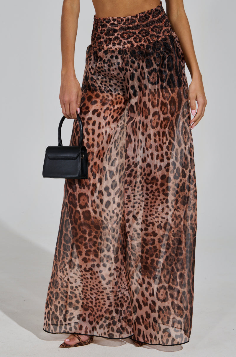 A person wearing the CHEETAH GIRL PALAZZO PANT, characterized by its high-waisted and wide-leg design in a bold cheetah print on sheer fabric, holds a small black handbag. They stand showcasing their midsection and part of their arms.