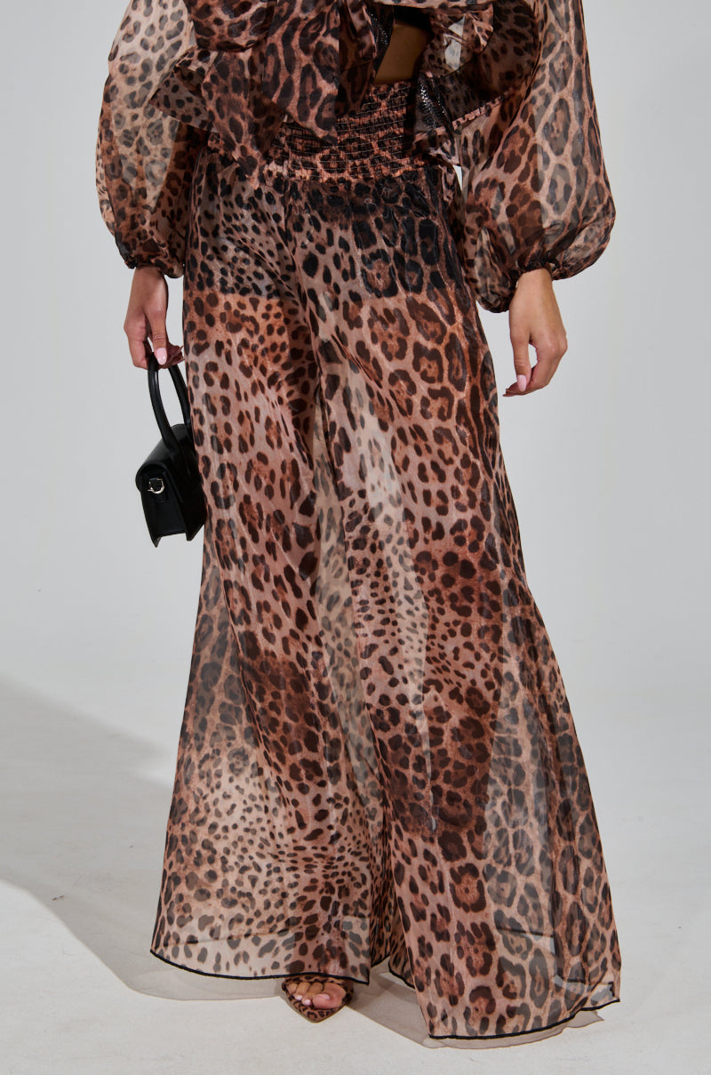 A person styled in the CHEETAH GIRL PALAZZO PANT, featuring a sheer, wide-legged cheetah print design, is paired with a matching blouse adorned with voluminous sleeves. They carry a small black bag, with their shoes subtly peeking out from beneath the high-rise fit pants against a plain, light-colored background.