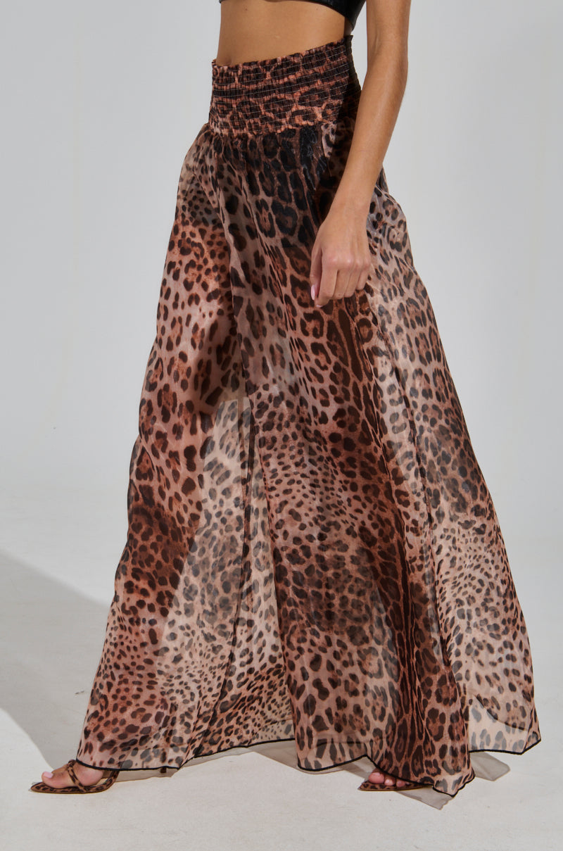 A person is wearing a pair of CHEETAH GIRL PALAZZO PANTS, which are flowy with a cheetah print. These sheer pants have a high-rise fit and feature a high, gathered waistband. The image captures the lower body, highlighting the design and movement of the pants paired with a black crop top.