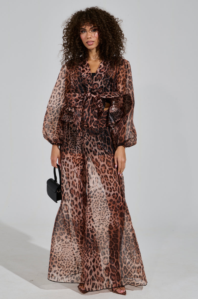 A person with curly hair wears the CHEETAH GIRL PALAZZO PANT, paired with a sheer, long-sleeve outfit and accessorized with a small black handbag against a plain white background.