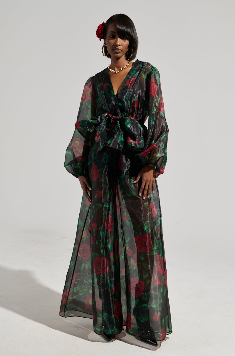 A person wearing the elegant "I Wish You Roses Palazzo Pant," featuring a flowing sheer fabric with rose prints in red and green, stands gracefully against a plain background. A red flower adorns their hair, enhancing the floral theme.