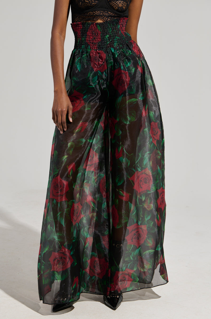 A person wearing the I WISH YOU ROSES PALAZZO PANT, which is sheer and high-waisted, showcasing a rose print with vibrant red roses and green leaves. The pants feature a smocked waistband and elegantly flow down to the ankles, with a neutral background accentuating the striking floral design.