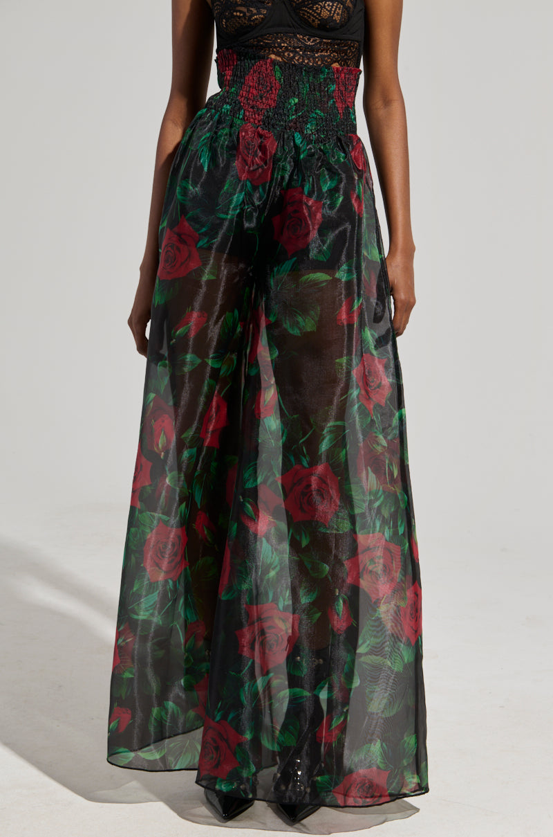 A person wearing the I WISH YOU ROSES PALAZZO PANT, featuring a sheer, floral maxi skirt in a stunning rose print with red roses and green leaves on a black background. The high-rise fit of the skirt gracefully complements the palazzo pants underneath, set against a plain backdrop.