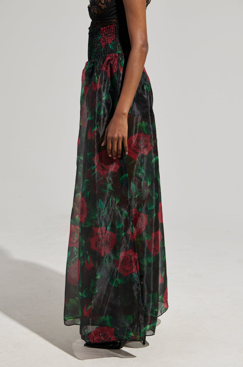 The individual is dressed in a high-rise fitted I Wish You Roses Palazzo Pant, adorned with a sheer overlay showcasing an elegant pattern of red roses. They stand against a simple and light-colored backdrop.