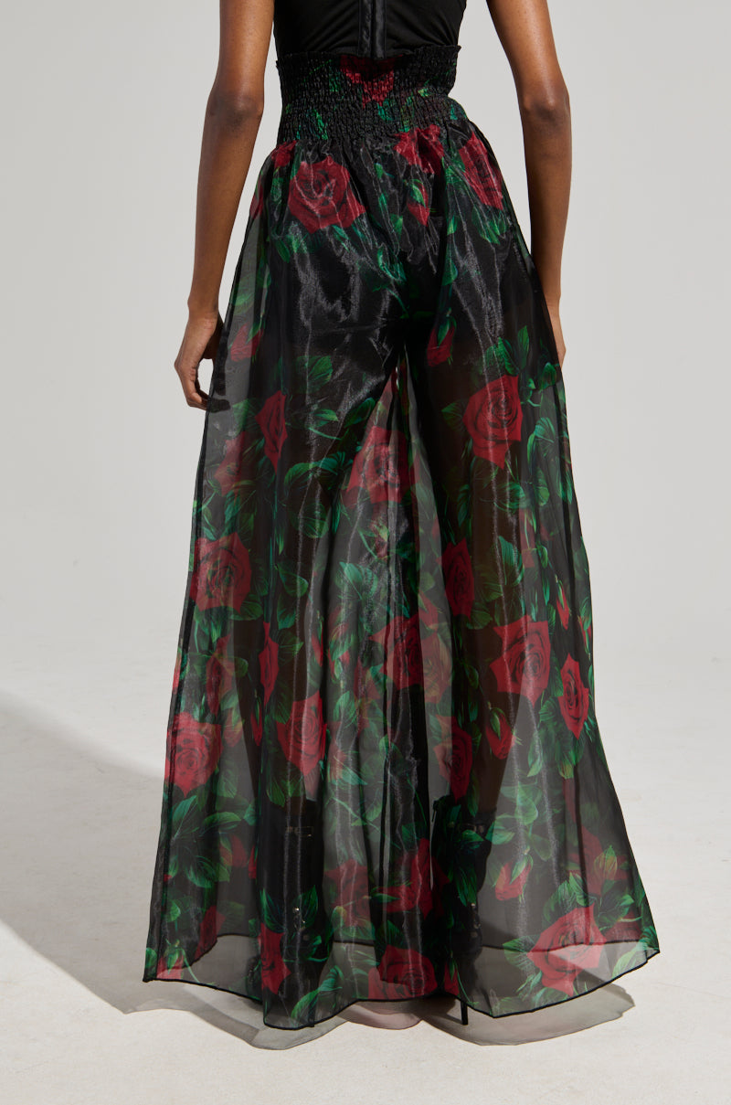 A person is wearing a black top combined with the I WISH YOU ROSES PALAZZO PANT, which boasts a long, flowing design with a sheer overlay decorated in rose print. The image captures the elegance of the back view, where each red rose detail enhances the charm of this ensemble.