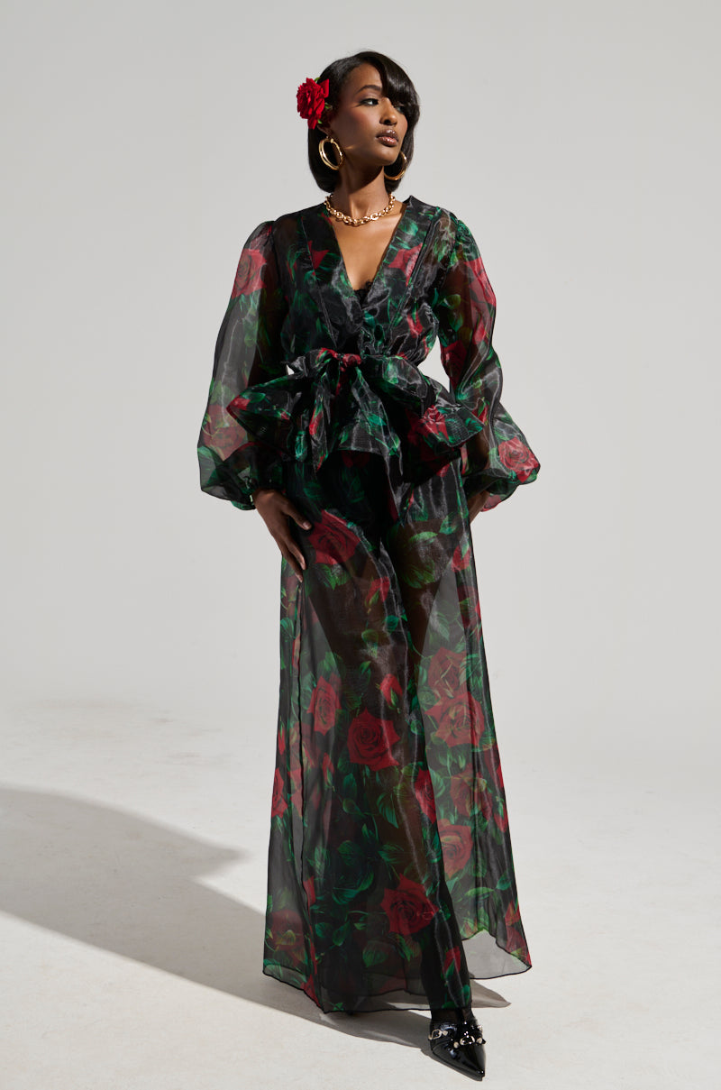 Wearing the elegant "I Wish You Roses Palazzo Pant," a woman stands against a plain background in a sheer black ensemble adorned with red rose patterns. She accessorizes her look with gold jewelry and completes it with a red flower in her hair.