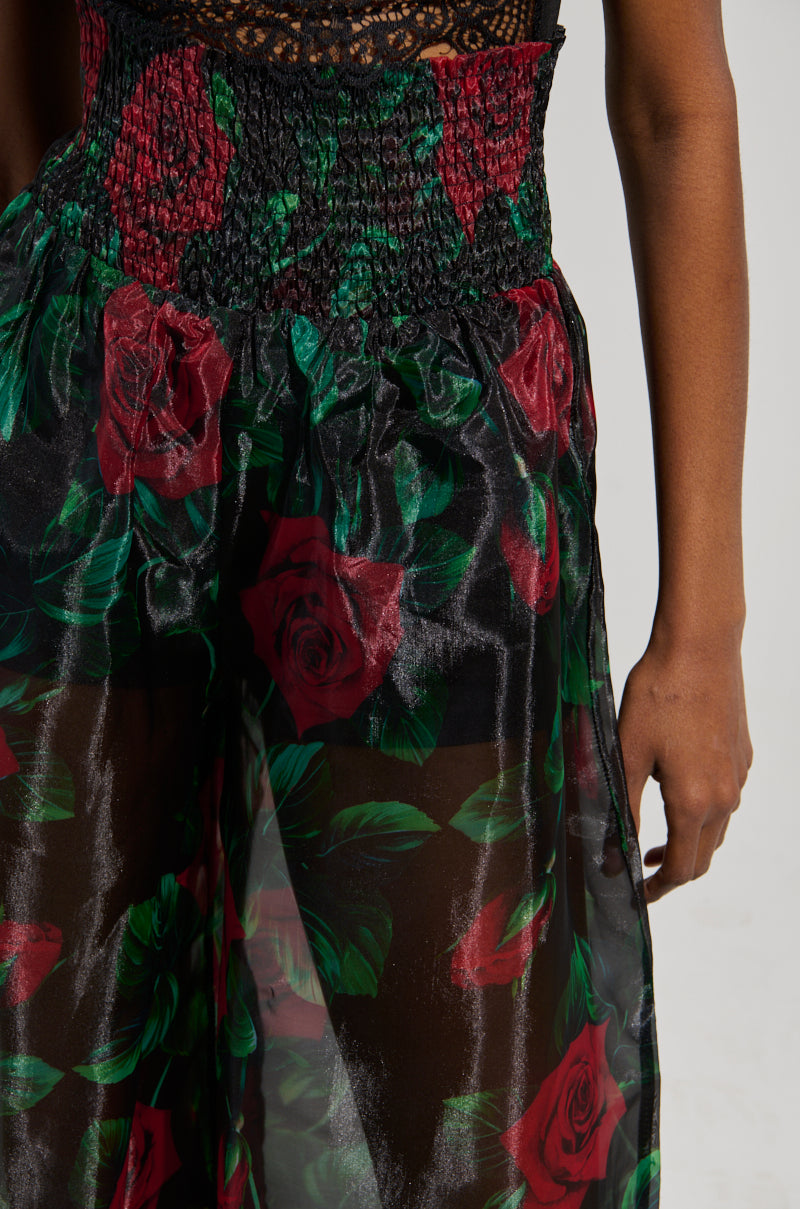 Close-up of a person wearing the I WISH YOU ROSES PALAZZO PANT in sheer black with a vibrant rose print. The high-rise fit is gathered at the waist, creating a textured look as they stand against a plain background, beautifully showcasing the floral design.
