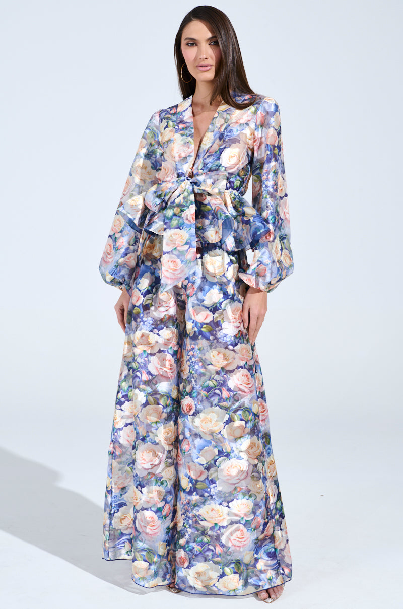 A woman in the EXALTED FLORAL PALAZZO PANT set, featuring a floral-patterned peplum top and high-rise, wide-legged pants in blue with pink, white, and yellow flowers, stands against a plain light background.