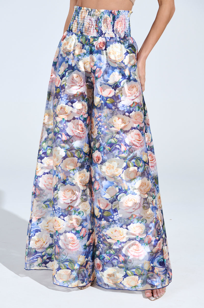 Someone showcases the EXALTED FLORAL PALAZZO PANT, featuring a high-waisted, wide-leg silhouette with colorful floral prints on sheer mesh. The pants have a smocked waistband and partially reveal the feet, indicating a flowy, lightweight fabric design.