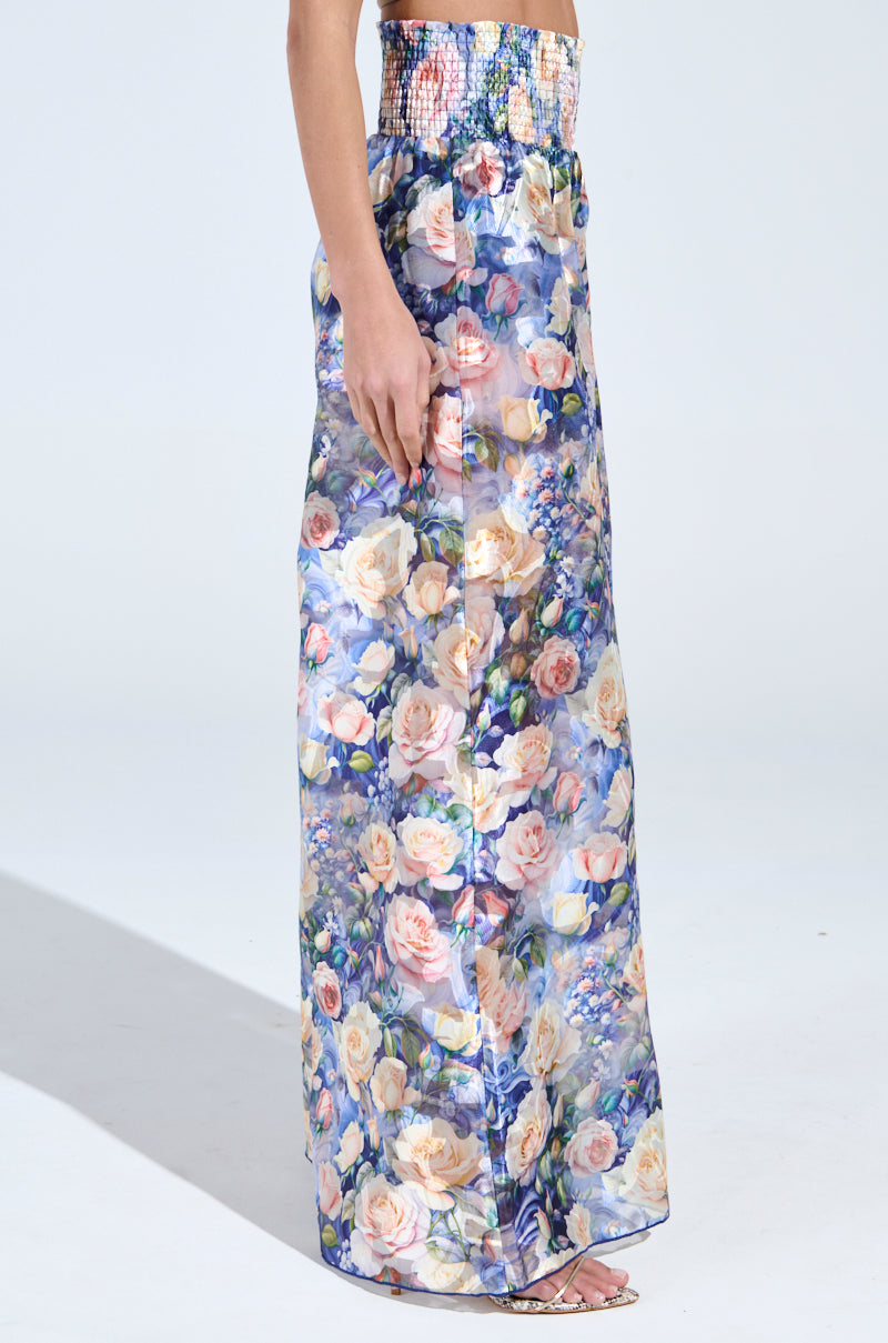 An individual wearing EXALTED FLORAL PALAZZO PANT stands against a light background. The sheer mesh overlay adds an ethereal touch to the vibrant roses and floral patterns, while the smocked colored waistband enhances its look. The outfit is completed with sandals.