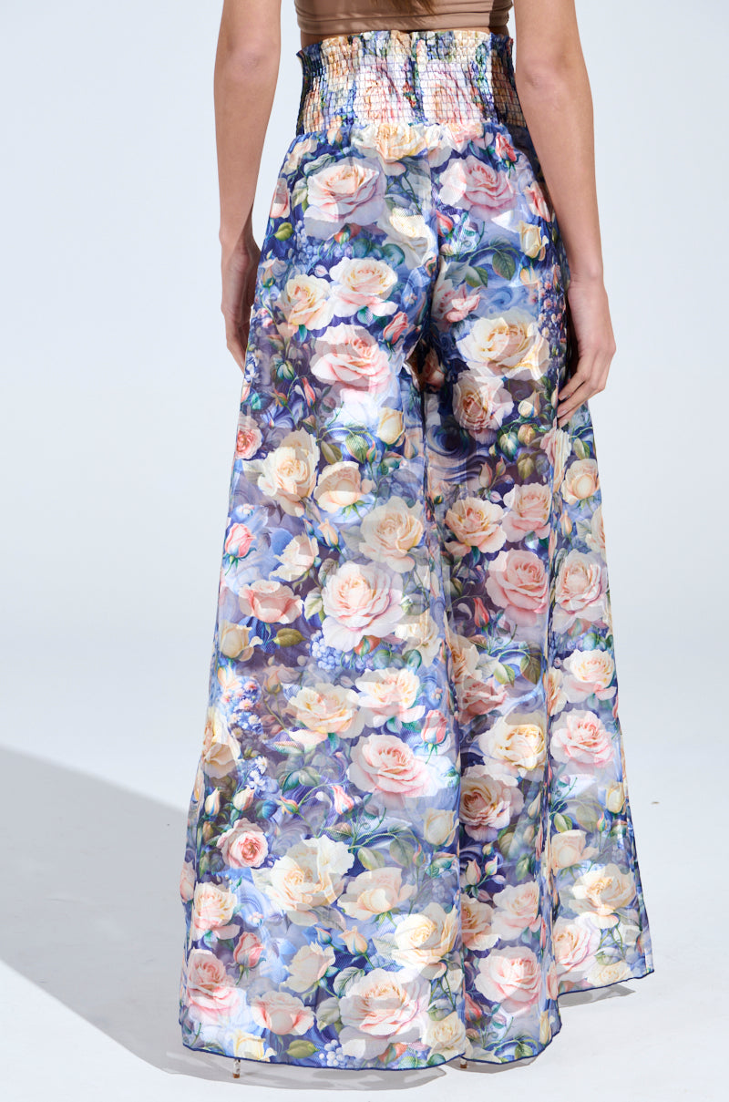 Someone is wearing the EXALTED FLORAL PALAZZO PANT, featuring a smocked high-rise fit with vibrant floral patterns of roses and leaves in pink, blue, and green shades against a light background.