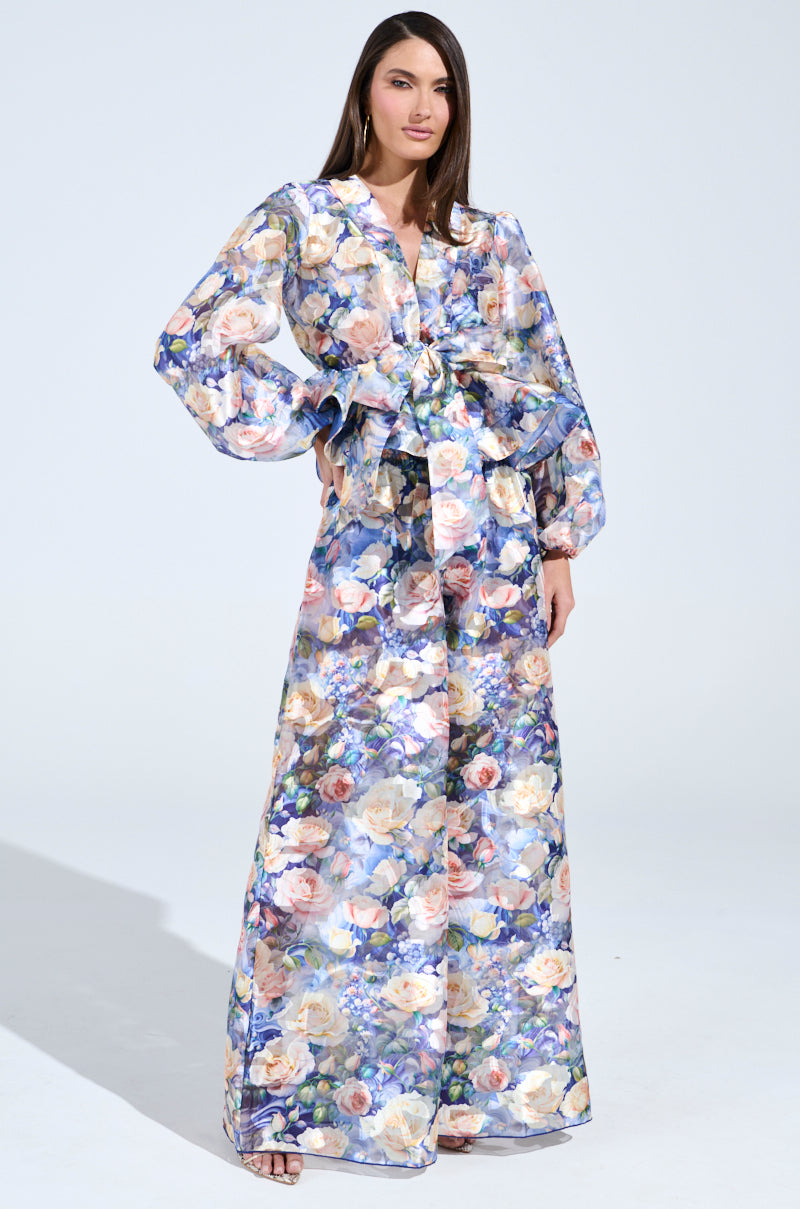 Wearing an EXALTED FLORAL PALAZZO PANT with a long, flowing dress in soft pastel colors and intricate floral patterns, sheer mesh sleeves, and tied waist detail, a person stands confidently against a plain white background.