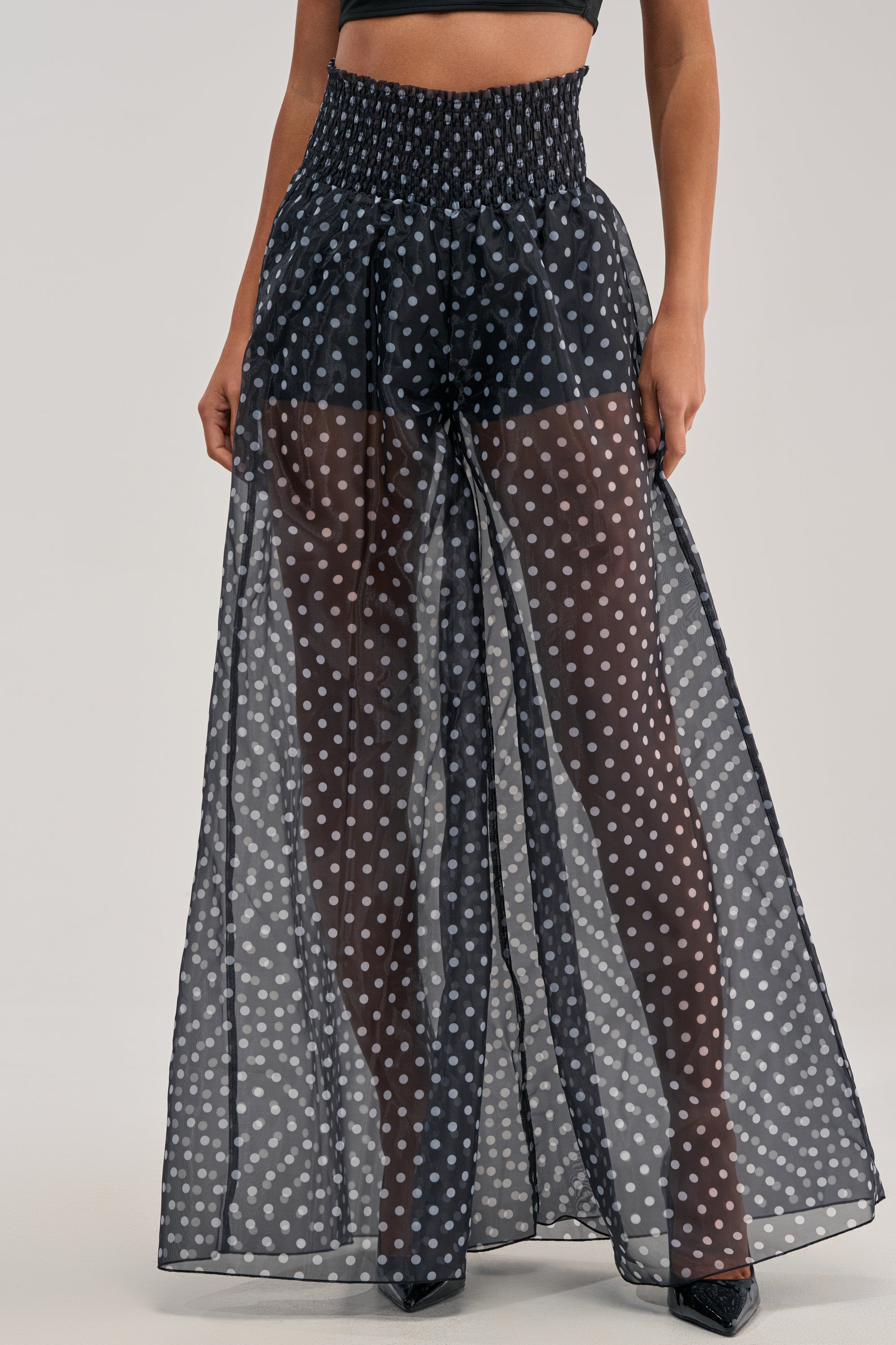Wearing the PRETTY GIRL POLKA DOT PALAZZO PANT, which features sheer high-rise chiffon with white polka dots over black shorts, styled with a sleek black crop top for a modern, chic appearance.