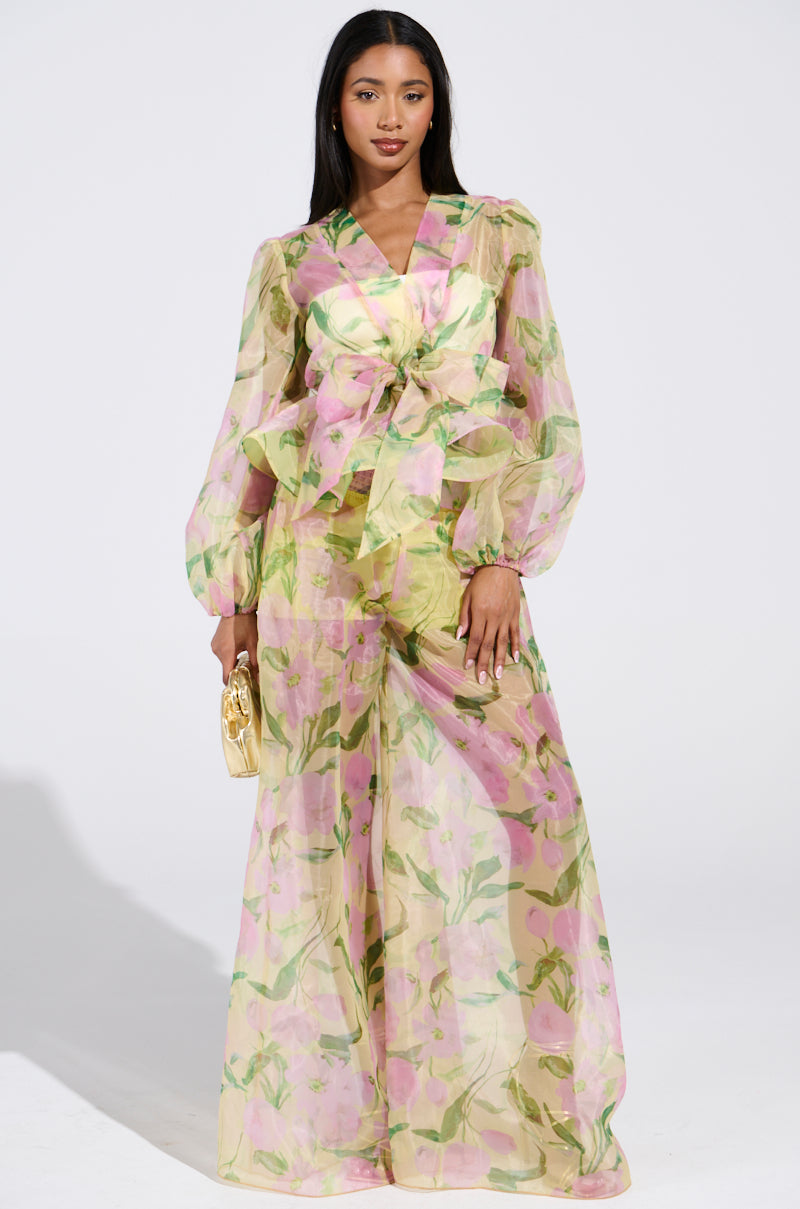 A woman exudes confidence in a floral outfit, donning a sheer long-sleeved top with a waist tie and Flower Power Wide Leg Palazzo Pants. Holding a small gold clutch, she effortlessly blends florals and bold style against the white backdrop.