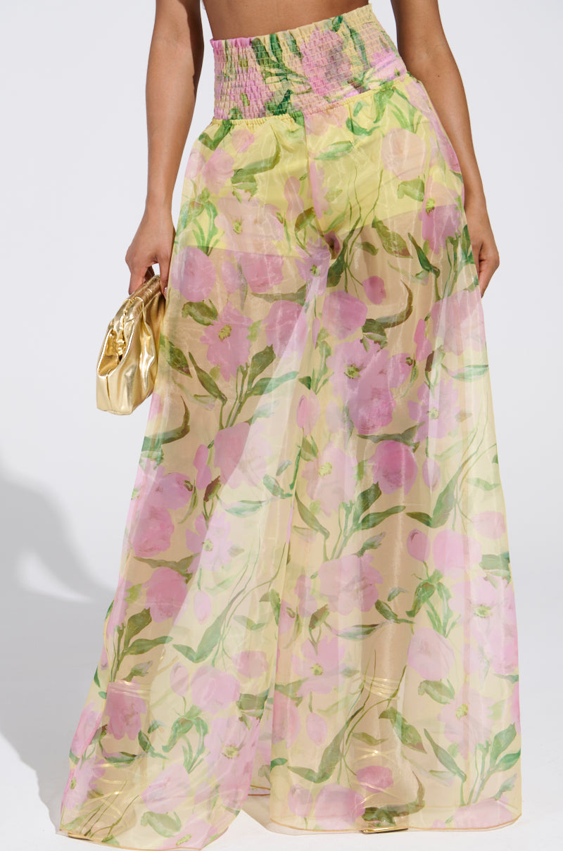 Wearing the FLOWER POWER WIDE LEG PALAZZO PANT, featuring a colorful floral pattern in pink, yellow, and green on chiffon fabric, the person holds a shiny gold clutch against a plain white background.
