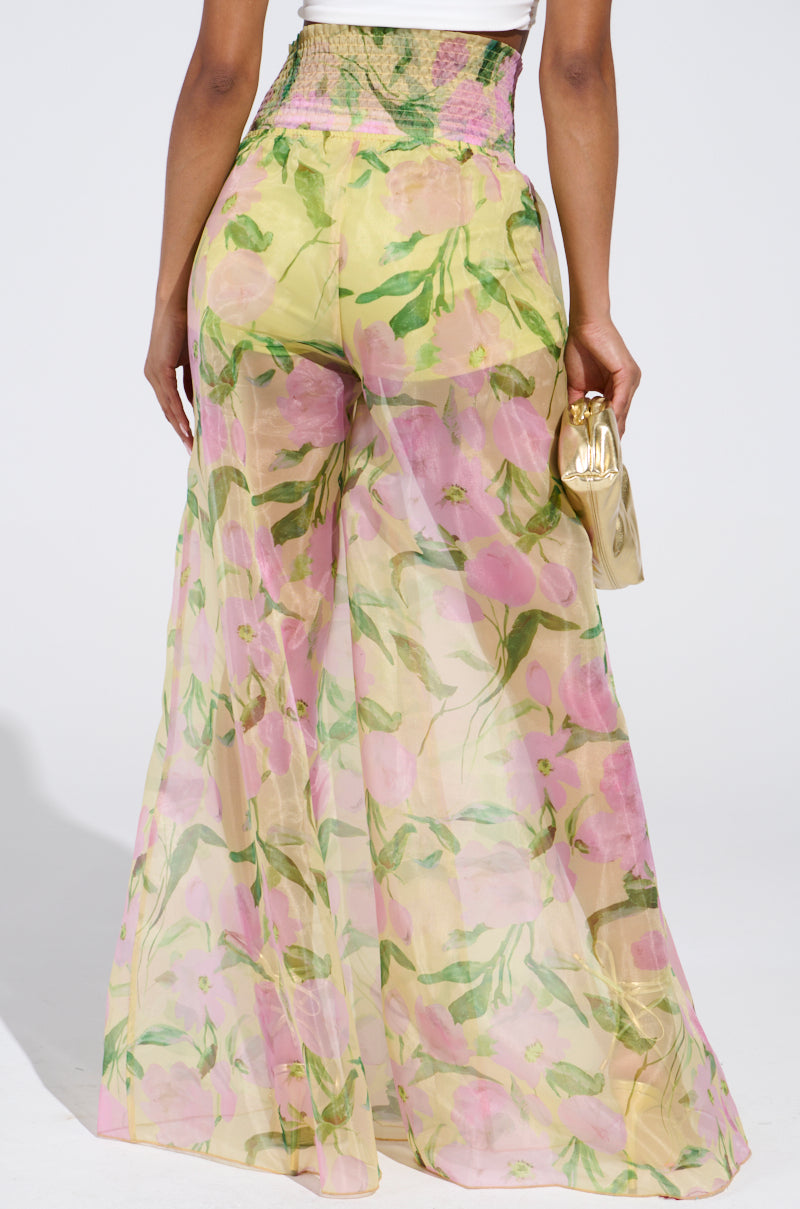 Elegantly dressed in high-waisted FLOWER POWER WIDE LEG PALAZZO PANT with a pink and green floral pattern, they carry a small gold clutch in their right hand against a simple, light backdrop.