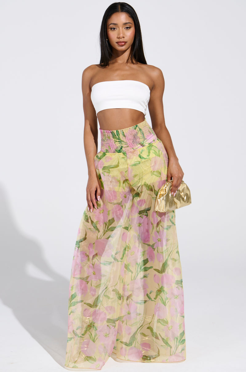 A woman confidently poses in a white strapless top and the sheer chiffon FLOWER POWER WIDE LEG PALAZZO PANT, adorned with pink and green patterns. She elegantly holds a small gold clutch, standing against a plain background.