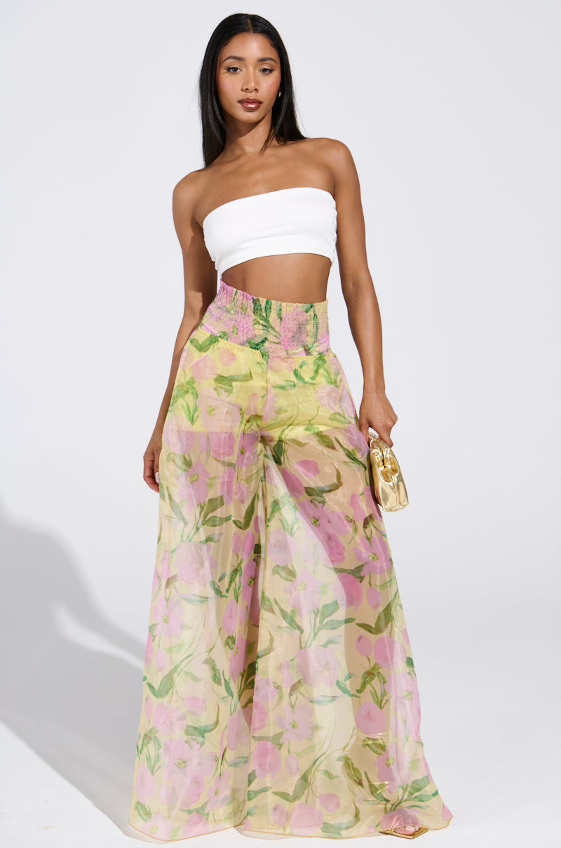 Against a plain backdrop, someone confidently models the FLOWER POWER WIDE LEG PALAZZO PANT—a pair of high-waisted, sheer floral pants—paired with a white tube top. They hold a small gold purse and have long, straight hair.