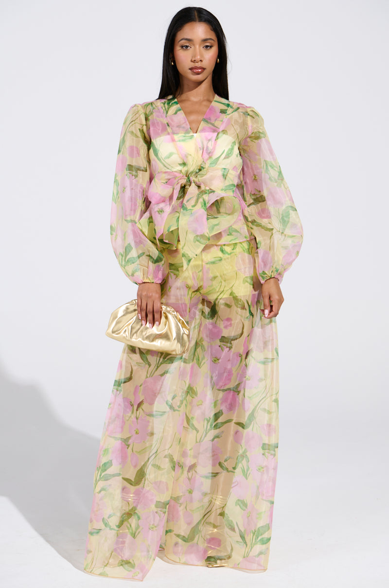 A woman with long, dark hair stands against a plain background wearing FLOWER POWER WIDE LEG PALAZZO PANTS with pink and green floral chiffon, voluminous legs, and a bow at the waist. She holds a small, gold clutch in one hand.